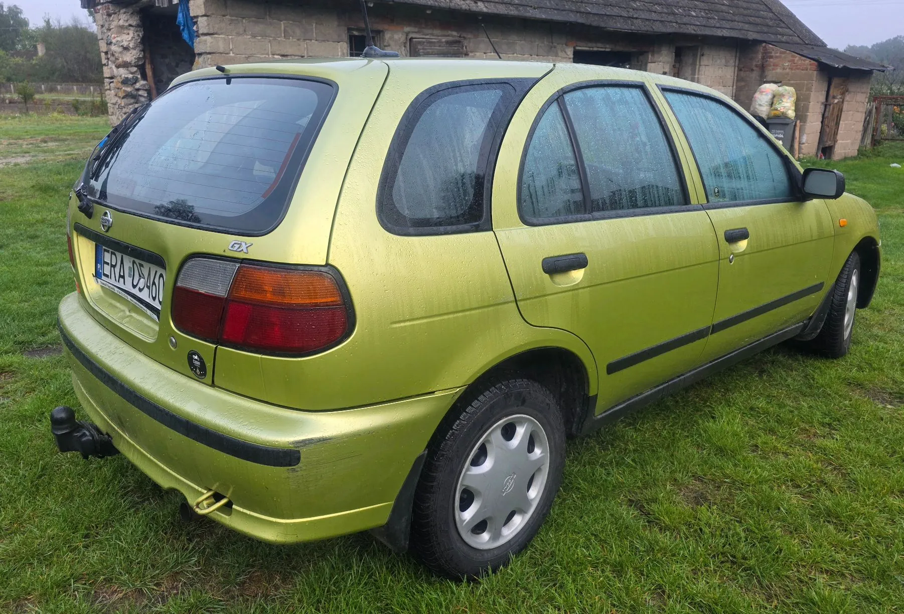 Jasnozielony samochód osobowy typu hatchback zaparkowany na trawie. Widoczny jest tył oraz prawy bok pojazdu. Samochód ma tablicę rejestracyjną ERA CD460 oraz oznaczenie modelu GX na klapie bagażnika. Na karoserii widoczne są krople wody. W tle znajduje się stary budynek z kamienia i cegły.