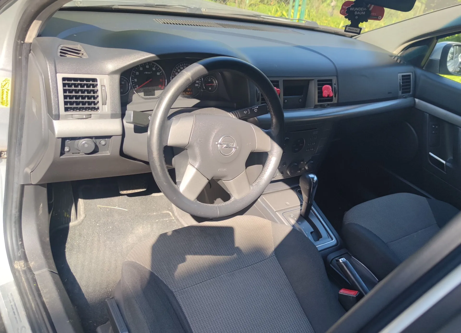 Wnętrze samochodu marki Opel, widok z perspektywy pasażera na stronę kierowcy. Widoczna jest czarna kierownica z logo producenta, deska rozdzielcza z analogowymi wskaźnikami prędkości i obrotów, kratki nawiewów oraz konsola środkowa z lewarkiem zmiany biegów i hamulcem ręcznym. W tle widoczne jest ciemnoszare, materiałowe siedzenie kierowcy.