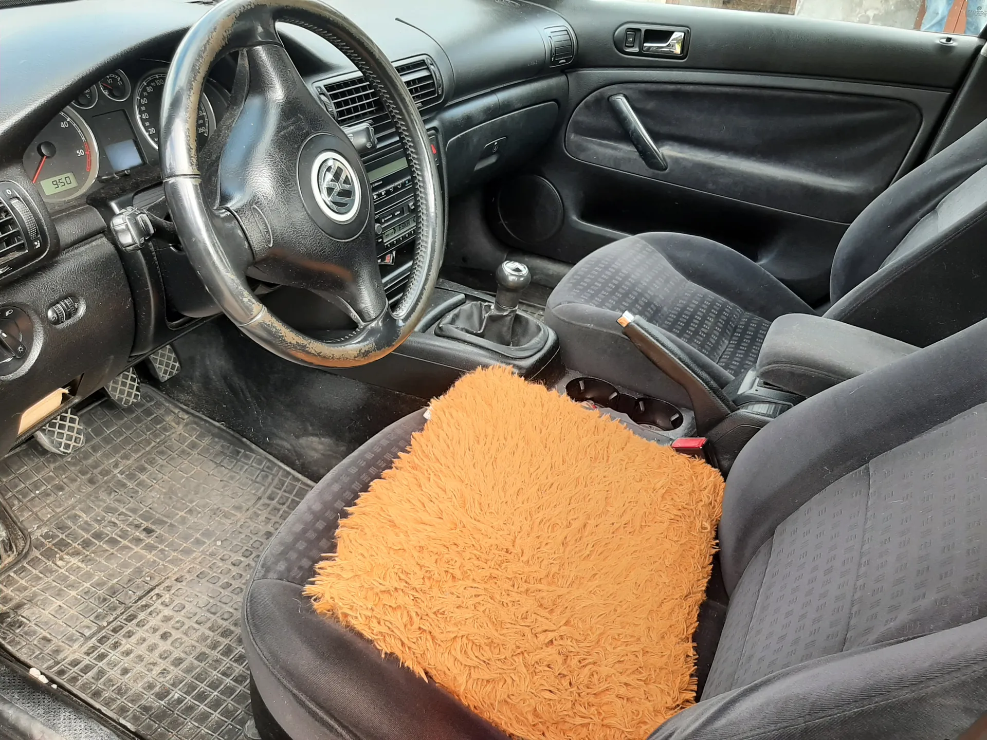 Wnętrze samochodu z widocznym kierownicą marki Volkswagen, czarnymi siedzeniami i pomarańczowym puszystym poduszką na siedzeniu kierowcy. Deska rozdzielcza z licznikami, w tym prędkościomierzem pokazującym 950, oraz centralnym systemem audio. Widoczny dźwignia zmiany biegów, drzwi z uchwytem i głośnikiem. Podłoga wyłożona czarnymi matami z siateczkowym wzorem. Kierownica i deska rozdzielcza mają ślady zużycia.