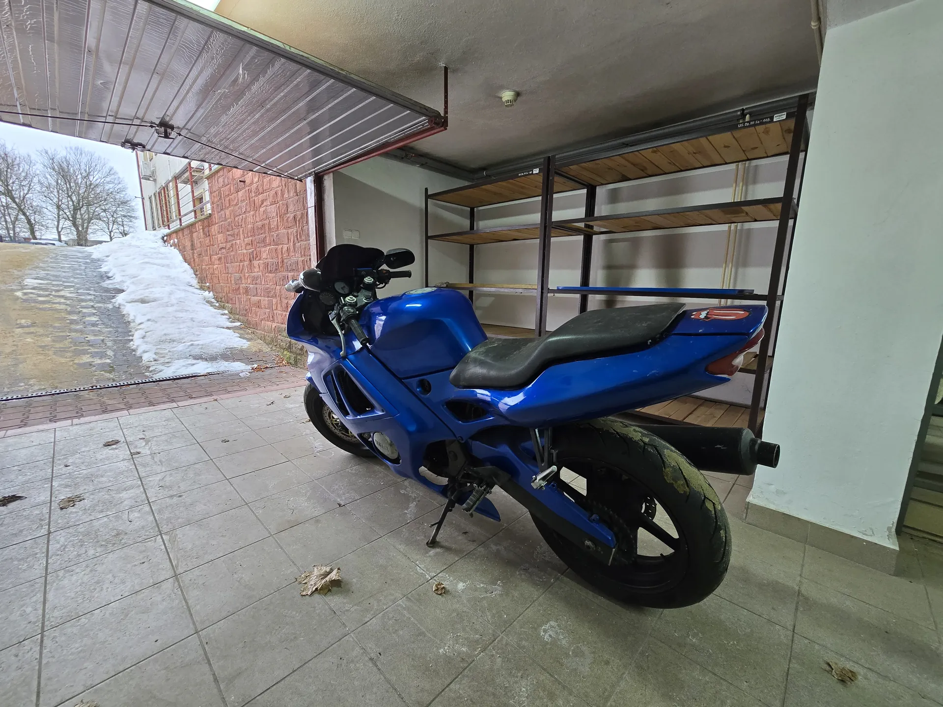 Niebieski motocykl sportowy z widocznym numerem "10" na tylnej części, zaparkowany pod daszkiem garażowym z metalowym dachem. W tle ceglana ściana, śnieg przy chodniku, bezlistne drzewa oraz puste metalowe półki. Podłoga wyłożona płytkami z rozrzuconymi suchymi liśćmi. Po lewej stronie widoczna droga z resztkami śniegu i budynki w oddali.
