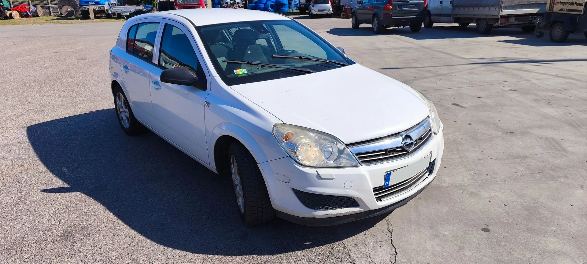 Biały samochód osobowy marki Opel Astra w nadwoziu hatchback, zaparkowany na betonowym placu. Auto jest widoczne z przodu i z boku. W tle znajdują się inne pojazdy, w tym samochody osobowe i przyczepy transportowe.