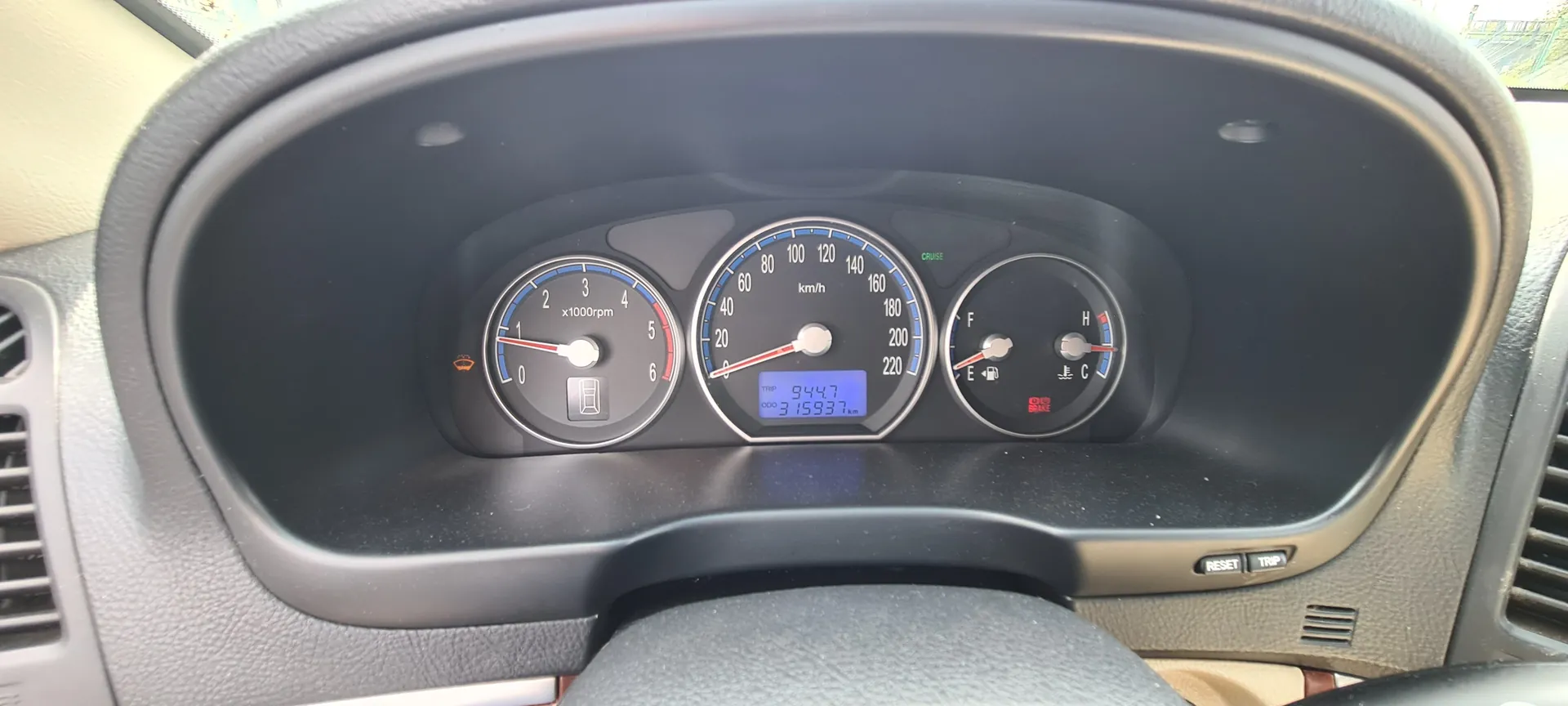 Zbliżenie na deski rozdzielcze samochodu z trzema wskaźnikami. Po lewej stronie wskaźnik obrotomieru z zakresem 0-6 (x1000rpm), na środku prędkościomierz w km/h z zakresem 0-220, a po prawej wskaźnik paliwa (blisko E) i temperatury chłodniczej (blisko C). W centralnym wyświetlaczu cyfrowym widoczne wartości: TRIP 944.7 i ODO 315937 km. Na lewym wskaźniku aktywny symbol ostrzegawczy (żarówka). Poniżej wskaźników przyciski oznaczone RESET i TRIP. W tle fragment kierownicy i deski rozdzielczej w kolorze szarym.