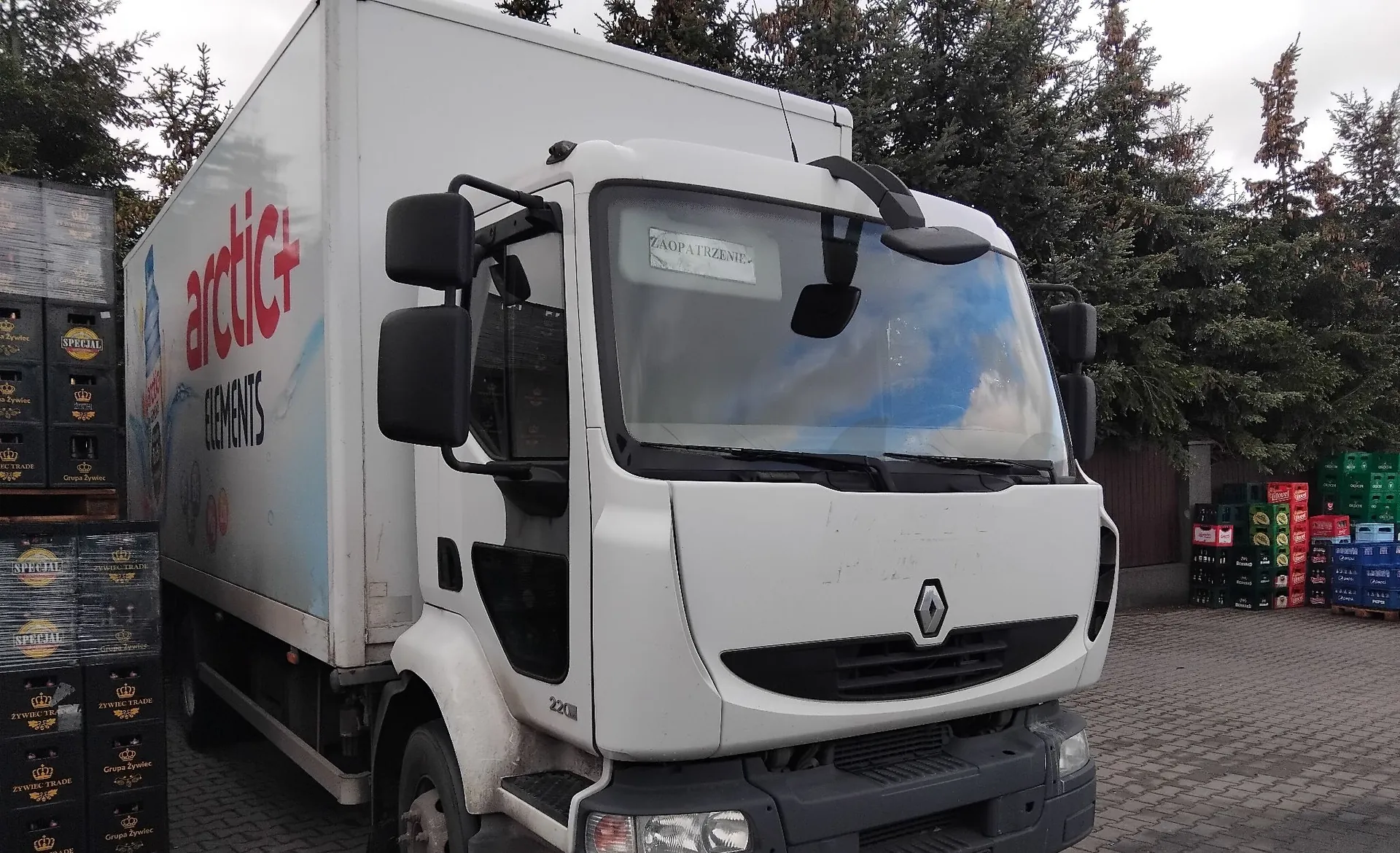 Biała ciężarówka marki Renault z zabudową chłodniczą. Na boku naczepy znajduje się niebiesko-czerwony napis „arctic+ ELEMENTS” oraz grafika z lodem i butelką napoju. W przedniej szybie kabiny umieszczona jest kartka z napisem „ZAOPATRZENIE”. Po lewej stronie widoczne są ułożone w stosy skrzynki z napojami, a w tle zielone drzewa iglaste.
