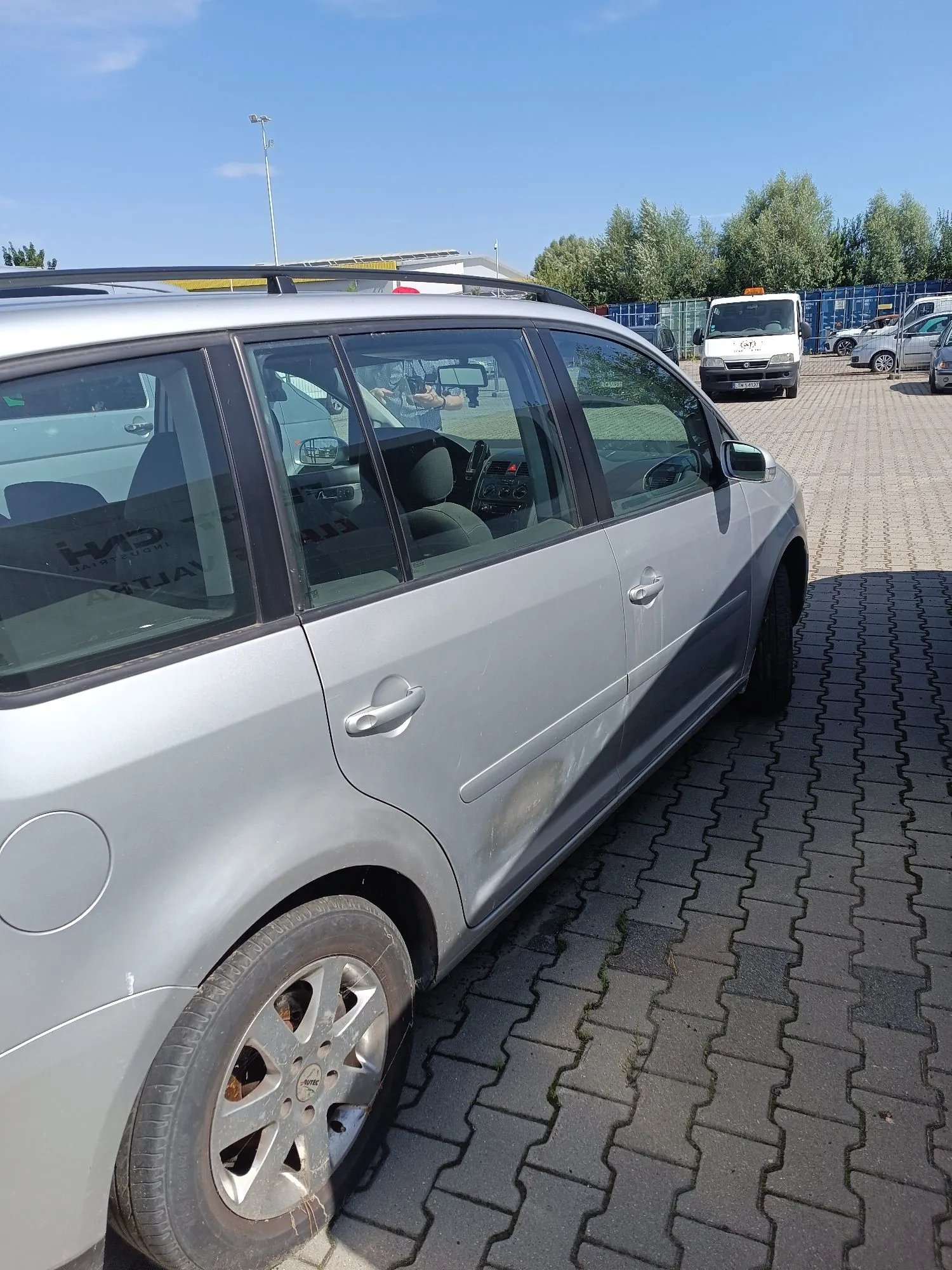 Srebrny samochód osobowy typu kombi zaparkowany na kostce brukowej. Zdjęcie przedstawia bok pojazdu, na którym widoczne są zarysowania i wgniecenia na drzwiach kierowcy oraz progu. W tle widać parking, biały samochód dostawczy oraz drzewa pod jasnoniebieskim niebem.
