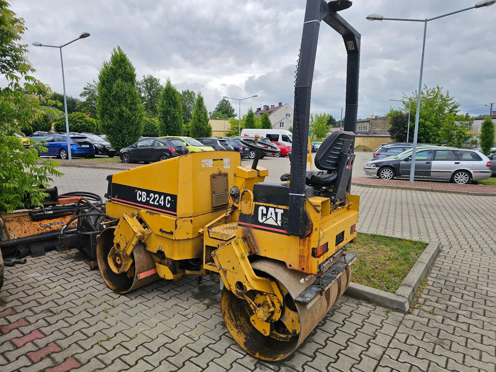 Żółty walec drogowy marki CAT, model CB-224 C, zaparkowany na kostce brukowej. Maszyna posiada dwa metalowe wałki i otwartą kabinę operatora z czarnym fotelem i stelażem ochronnym. W tle widoczny jest parking z zaparkowanymi samochodami osobowymi, zielone drzewa oraz jasne, zachmurzone niebo.