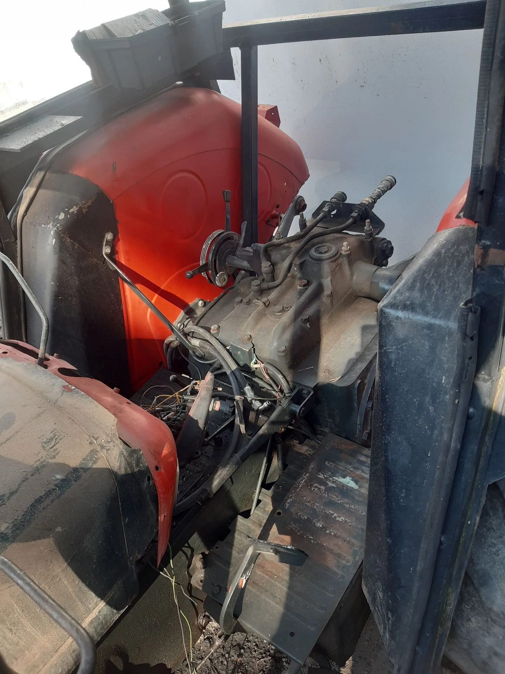 Zbliżenie na mechanizmy wewnętrzne i podzespoły maszyny rolniczej lub przemysłowej. Widoczna jest czerwona obudowa silnika, szare odlewy metalowe skrzyni biegów lub przekładni, liczne przewody, rurki oraz dźwignie sterujące. Całość znajduje się wewnątrz ciemnej, metalowej konstrukcji ramy.