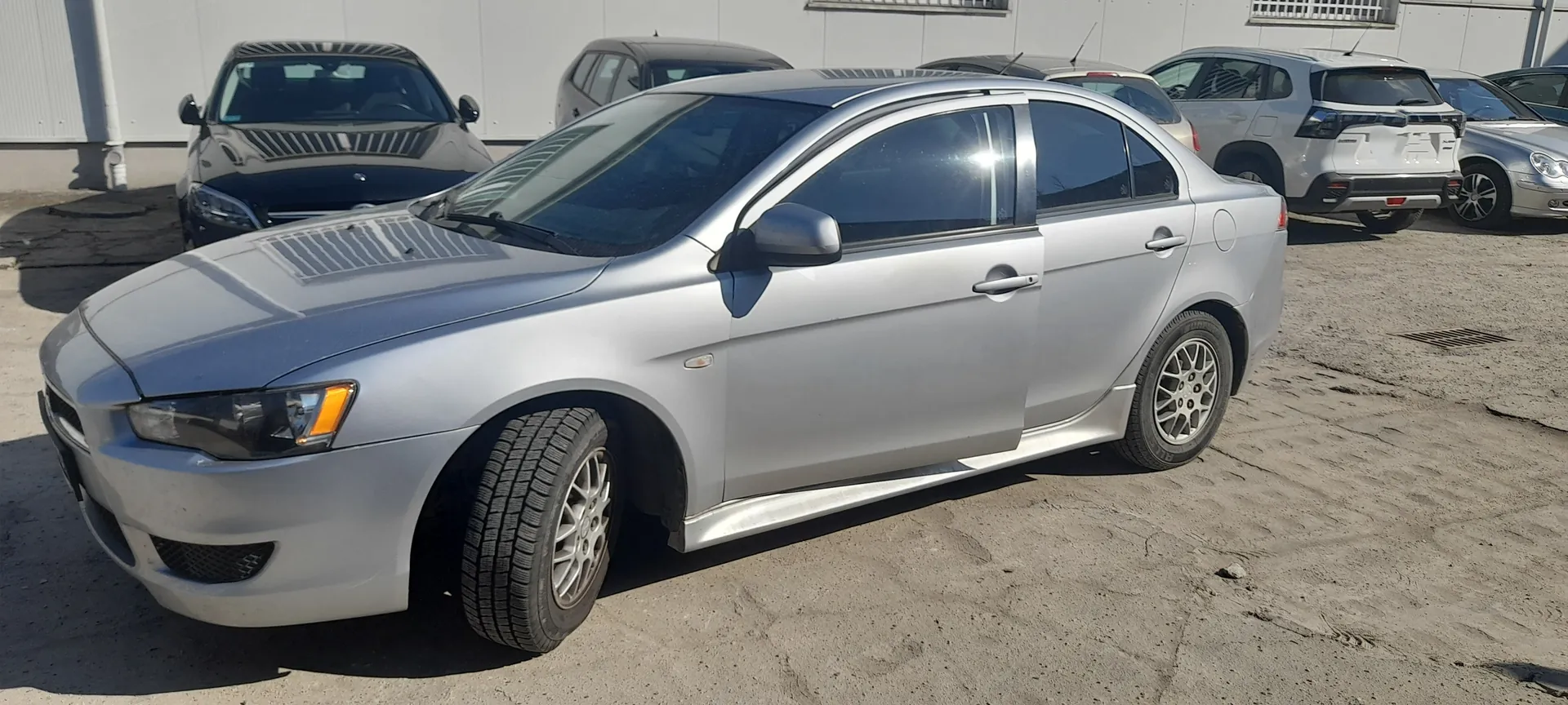 Srebrny samochód osobowy typu sedan na pierwszym planie, widoczny z boku i przodu. Pojazd ma cztery drzwi, srebrne felgi i dach z panoramicznym szybą. W tle stoją inne samochody: czarny sedan, biały SUV oraz inne pojazdy. Tło stanowi budynek z białymi ścianami i oknami. Samochody zaparkowane są na nierównym, piaszczystym terenie z widocznymi plamami asfaltu.