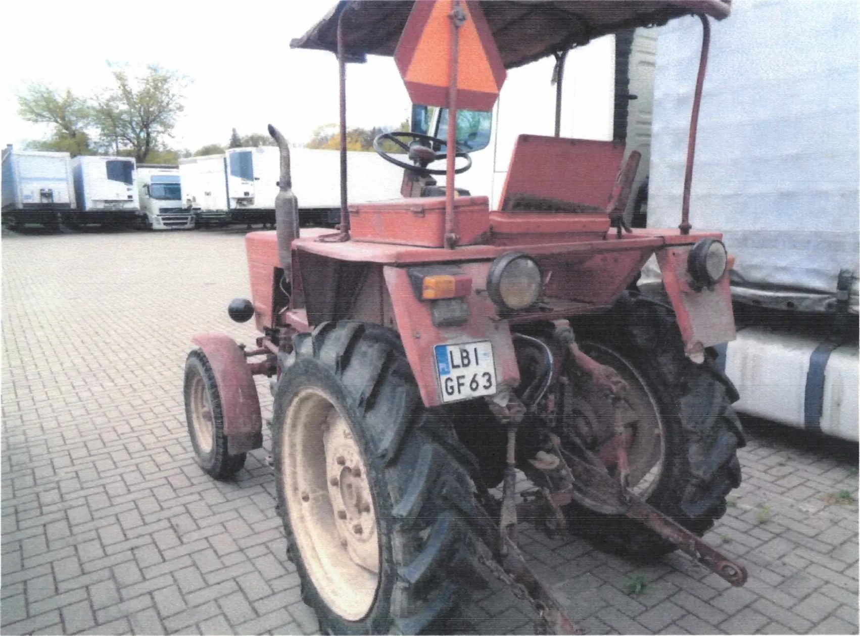 Czerwony traktor z tablicą rejestracyjną LBI GF63, stojący na brukowanej powierzchni. W tle widoczne są białe ciężarówki i drzewa. Traktor posiada pomarańczowy trójkątny znak ostrzegawczy oraz duże opony.