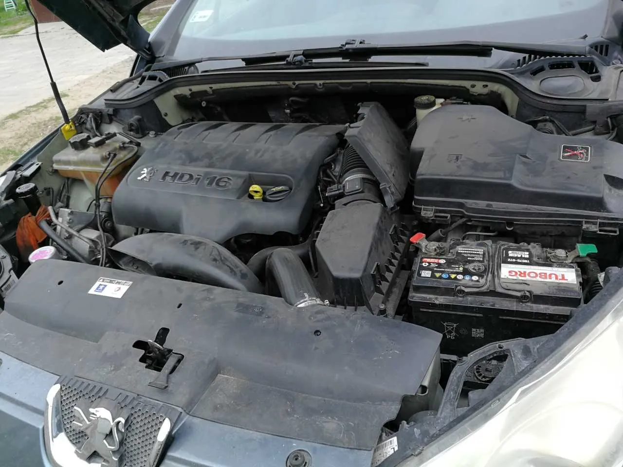 Otwarty kapota samochodu marki Peugeot z widocznym silnikiem oznaczonym HDi 16. W komorze silnika widoczne są elementy takie jak akumulator z napisem TUBORG, zbiornik płynu chłodniczego oraz różne przewody i osłony. Na przedniej części pojazdu znajduje się emblemat Peugeota. Tło przedstawia nierówną, żwirową powierzchnię oraz fragment drogi.