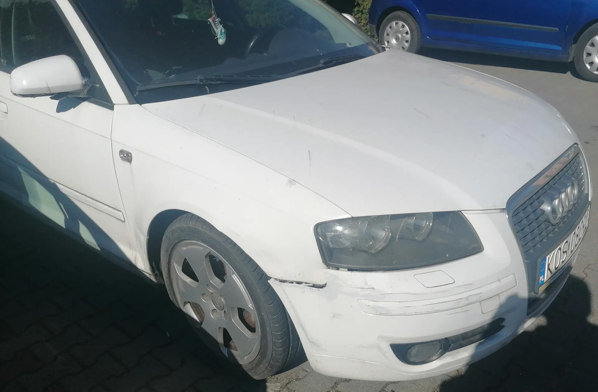 Biały samochód marki Audi z widocznym uszkodzeniem na przednim zderzaku i lewym reflektorze. Na masce widoczne są drobne zadrapania. Tablica rejestracyjna z napisem KOS 050H oraz oznaczeniem PL. W tle niebieski samochód zaparkowany na brukowanej powierzchni. Na przedniej szybie wiszą ozdoby.