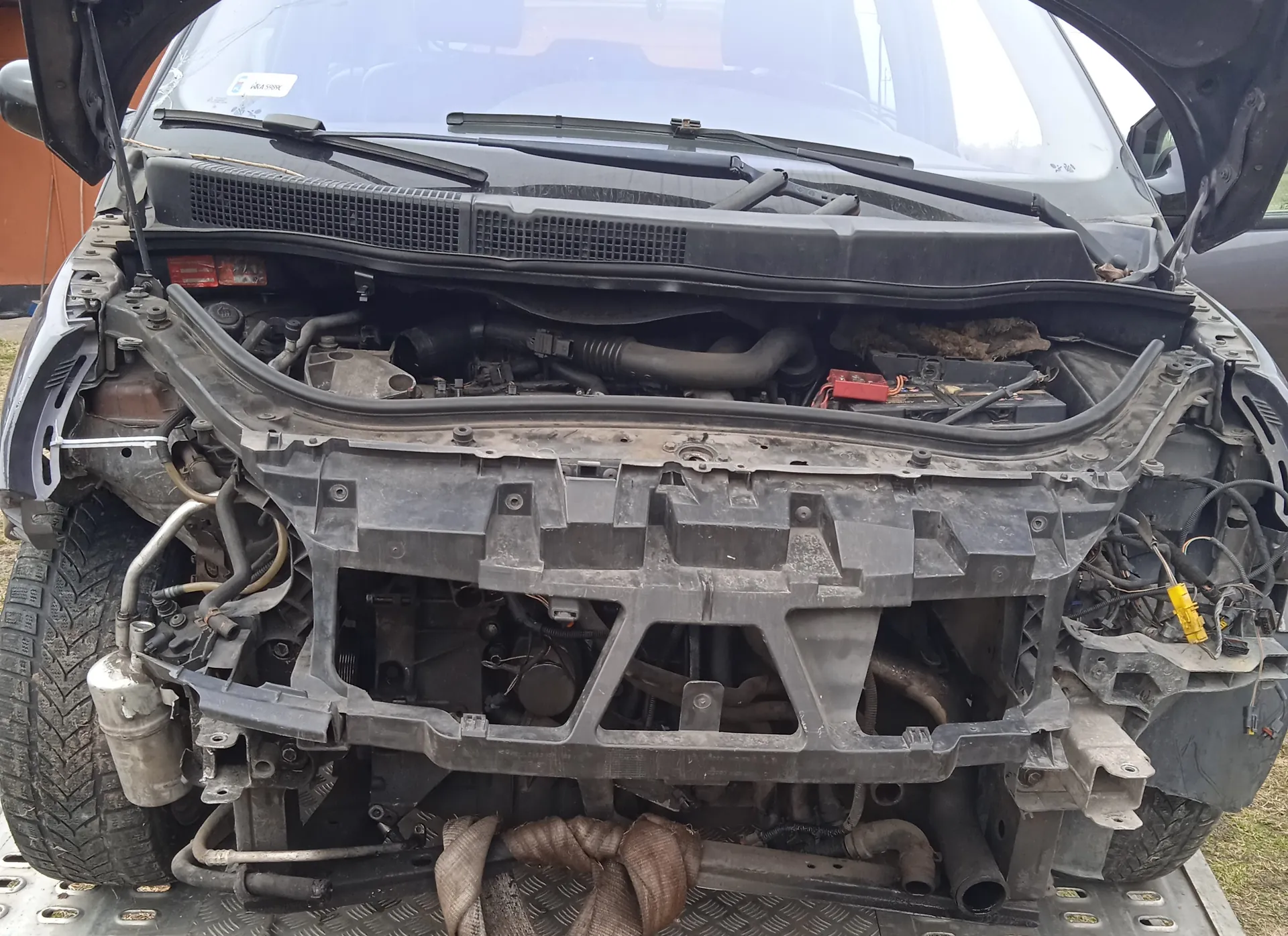 Czarny samochód z otwartą maską, ukazujący eksponowany silnik i elementy pod maską. Brakuje przedniego zderzaka, widoczne są odsłonięte przewody, rury oraz czerwony i żółty element elektryczny. Samochód stoi na metalowej platformie, z widocznymi oponami i fragmentem trawy oraz pomarańczową ścianą budynku w tle.