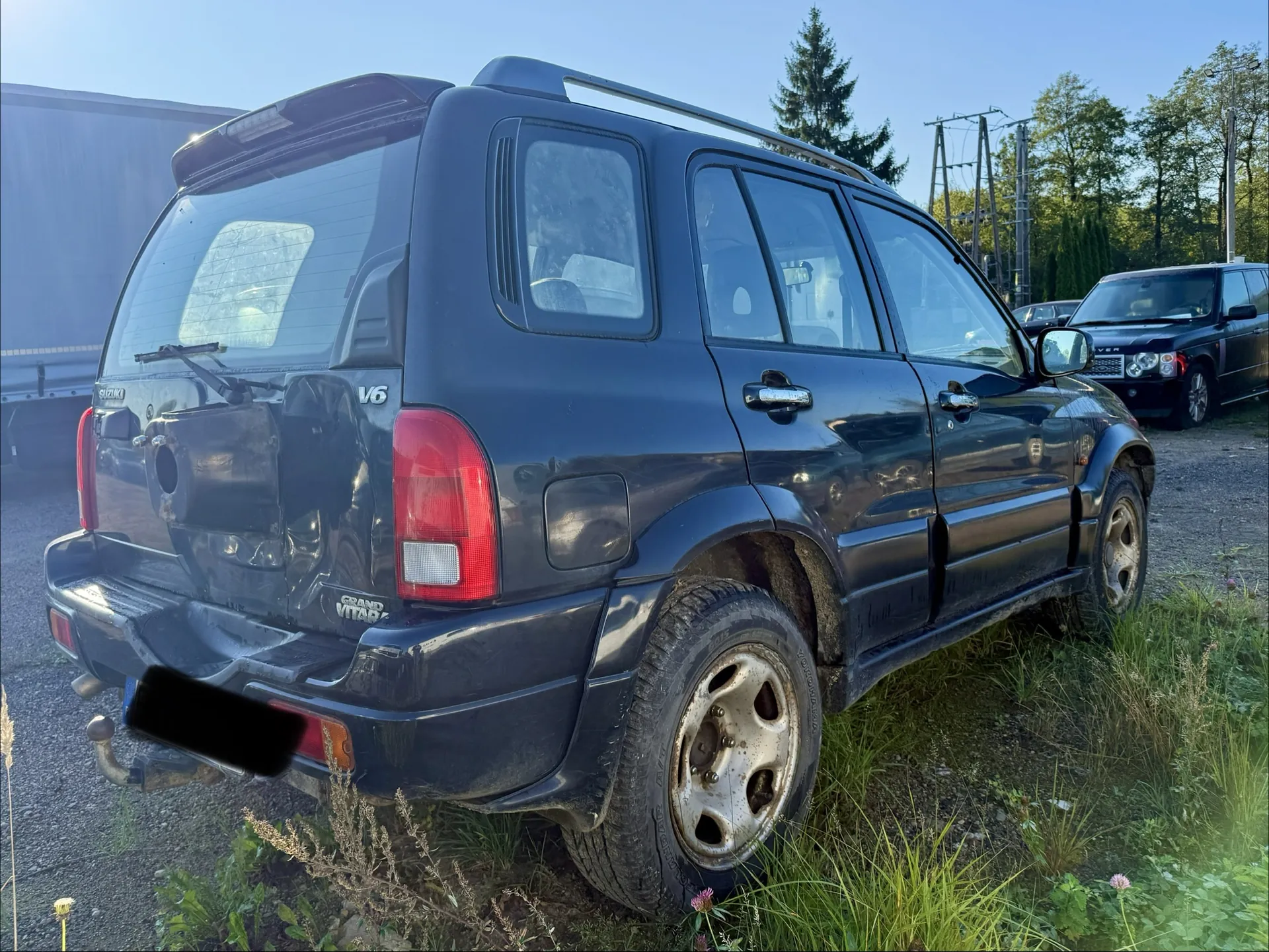 Czarny samochód terenowy marki Suzuki Grand Vitara V6, zaparkowany tyłem do obiektywu na trawiastym poboczu. Pojazd posiada relingi dachowe, tylny wycieraczki oraz widoczne napisy 'V6' i 'GRAND VITARA' na klapie bagażnika. Tablica rejestracyjna jest zakryta czarnym prostokątem. W tle widoczny jest inny czarny samochód oraz drzewa.