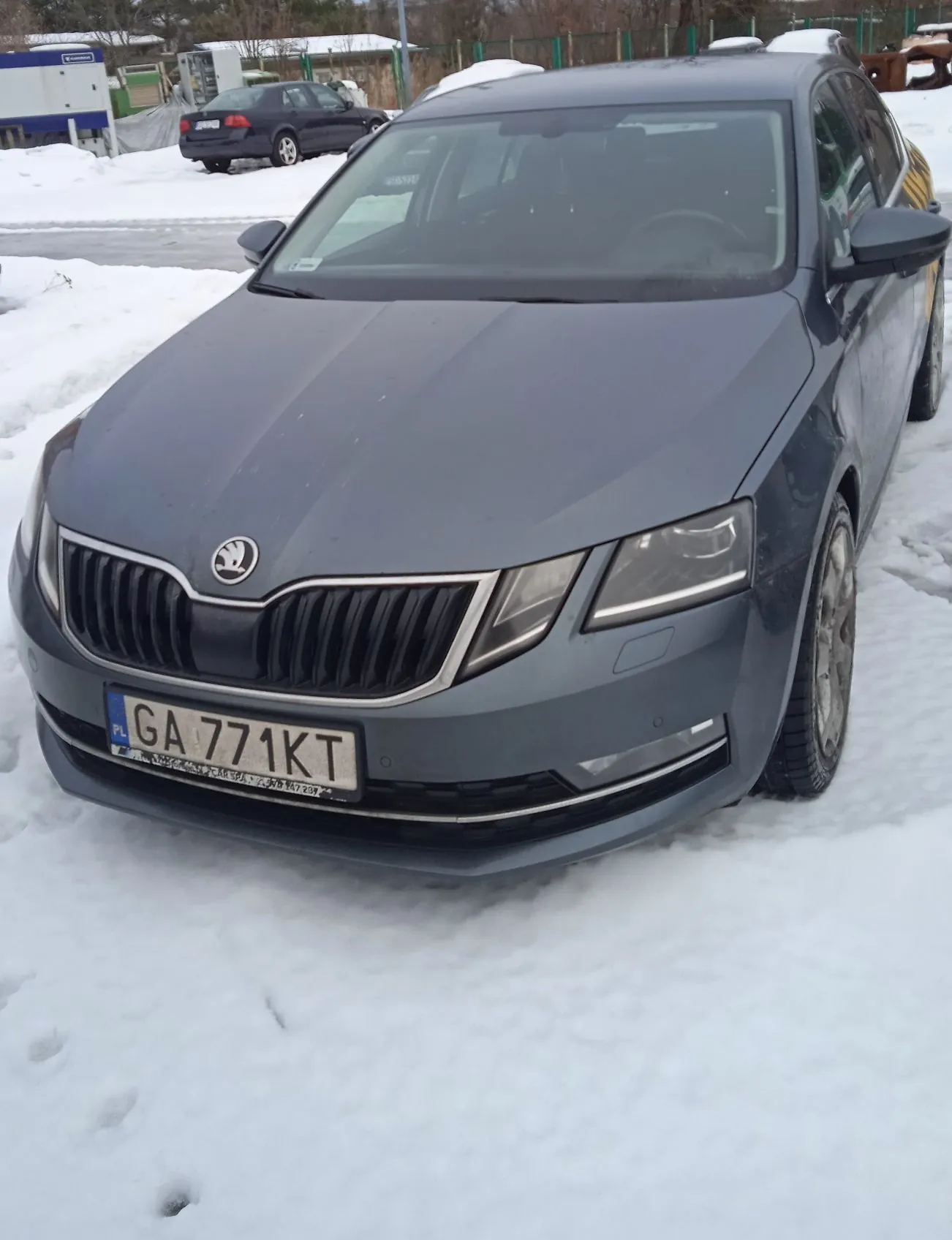 Szary samochód marki Škoda zaparkowany na śniegu. Widoczny jest przód pojazdu z tablicą rejestracyjną o numerze GA 771KT. W tle widać inny ciemny samochód oraz fragmenty budynków i ogrodzenia.