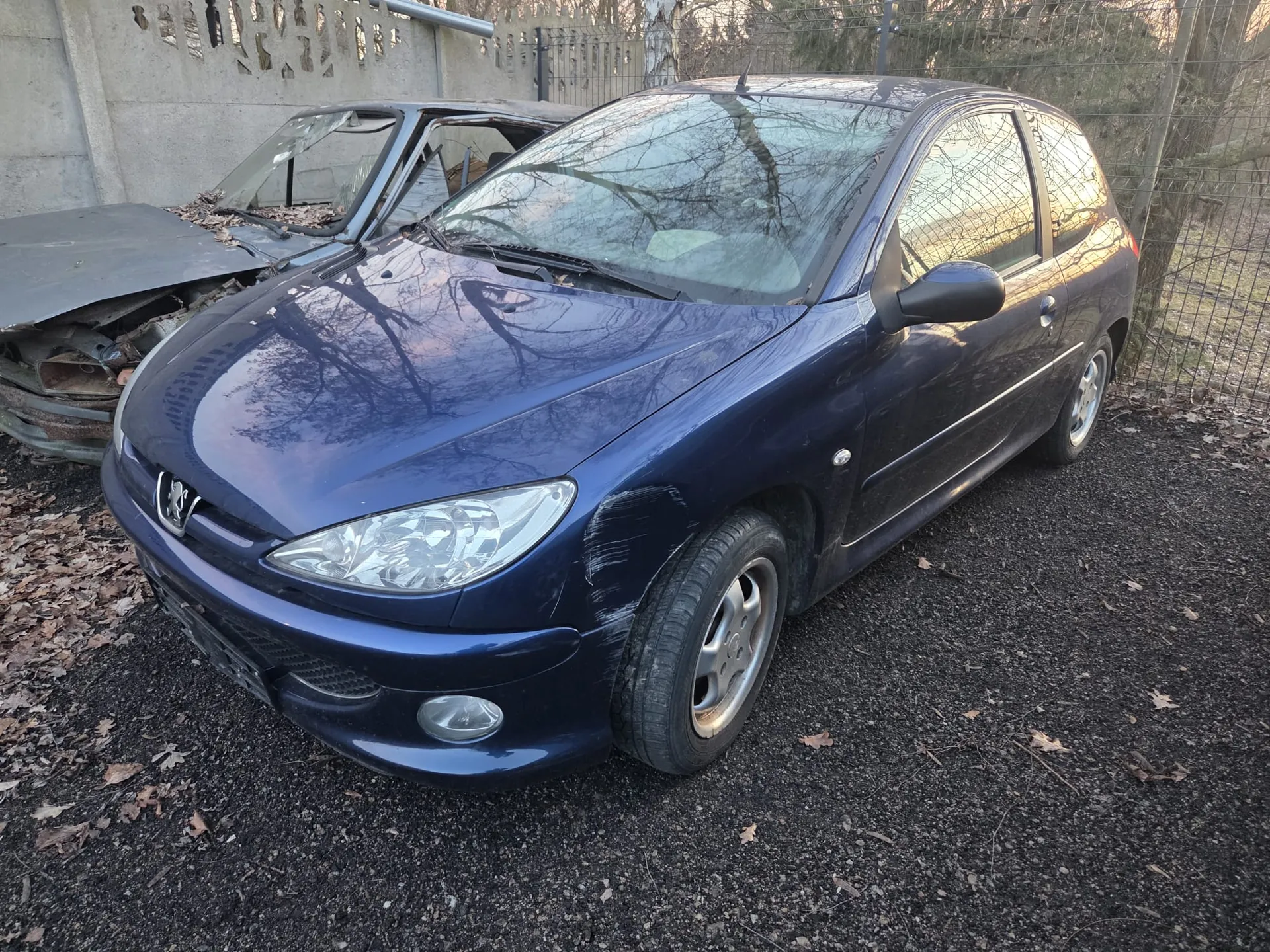 Ciemnoniebieski samochód osobowy marki Peugeot, trzydrzwiowy hatchback, zaparkowany na podłożu z drobnej kostki z rozsypanymi suchymi liśćmi. W tle uszkodzony samochód z otwartą maską i uszkodzonym przednim szybą, betonowa ściana z ozdobnymi wycięciami oraz metalowe ogrodzenie. W oddali widoczne drzewa bez liści i fragment trawy.