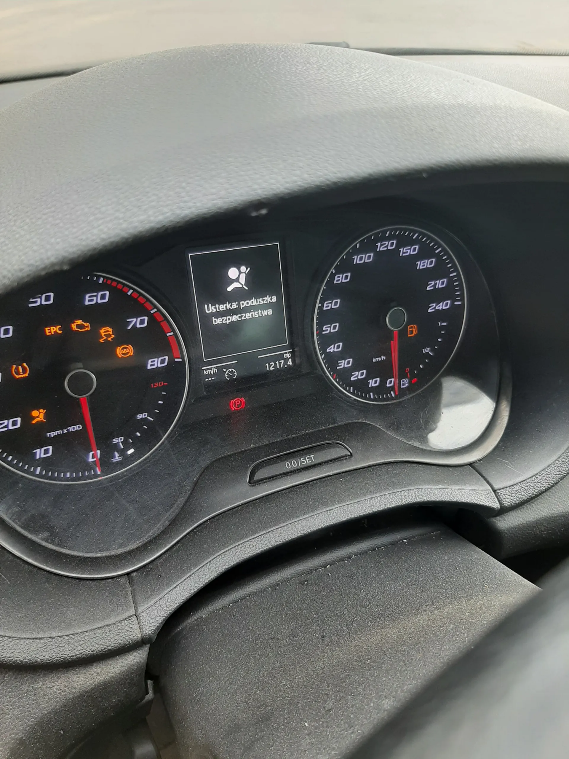 Zbliżenie na deski rozdzielcze samochodu. W górnej części znajduje się prędkościomierz z zakresem 0-240 km/h, wskazówka na 0. Poniżej wyświetlacz cyfrowy z napisem "Usterka: poduszka bezpieczeństwa" oraz ikoną poduszki powietrznej. Obok wyświetlacz z czasem 12:17.4 i czerwonym symbolem hamulca ręcznego. W dolnej części tachometr z zakresem 0-8000 obr./min, wskazówka na 0, oraz pomarańczowe lampki ostrzegawcze (m.in. EPC, kontrola silnika). Po prawej stronie przycisk oznaczony "0.0/SET". Wszystkie elementy są czarno-szare z czerwonymi i pomarańczowymi akcentami.