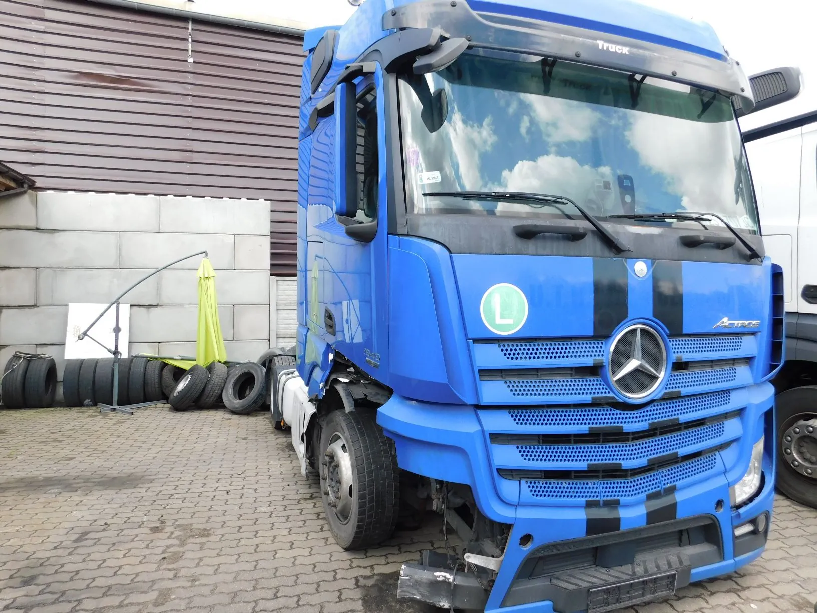 Niebieska ciężarówka marki Mercedes-Benz Actros z czarnymi pasami na masce i grillu. Pojazd posiada widoczne uszkodzenia w dolnej części z przodu oraz po prawej stronie kabiny. Na przedniej części kabiny znajduje się zielona okrągła naklejka z literą 'L'. Ciężarówka zaparkowana jest na kostce brukowej obok białego muru i brązowego budynku z blachy falistej. W tle widoczne są ułożone poziomo czarne opony.