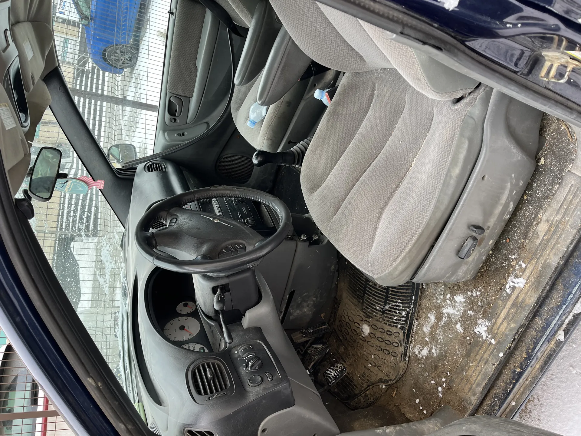 Wnętrze samochodu z otwartą drzwiami i lusterkiem bocznym. Szare tkaninowe fotele, kierownica, deska rozdzielcza z licznikami i przełącznikami klimatyzacji, dźwignia biegów. Butelka wody na fotelu pasażera. Podłoga i dywanik zabrudzone, śnieg na dywaniku. Widok na niebieski samochód i siatkę za oknem.
