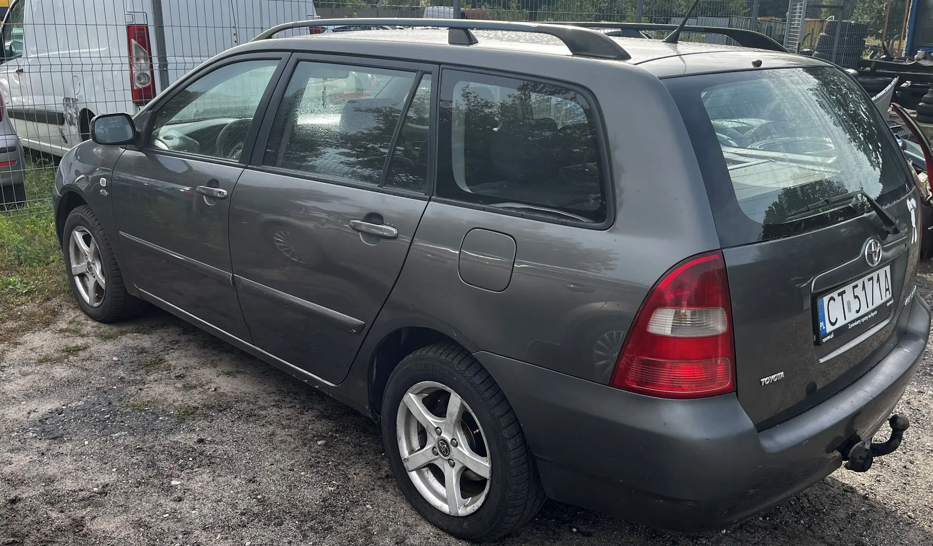 Szary samochód osobowy marki Toyota w nadwoziu kombi, zaparkowany na żwirowym podłożu. Widoczny jest tył i lewy bok pojazdu. Samochód posiada relingi dachowe oraz hak holowniczy. Tablica rejestracyjna ma numer CT 5171A.