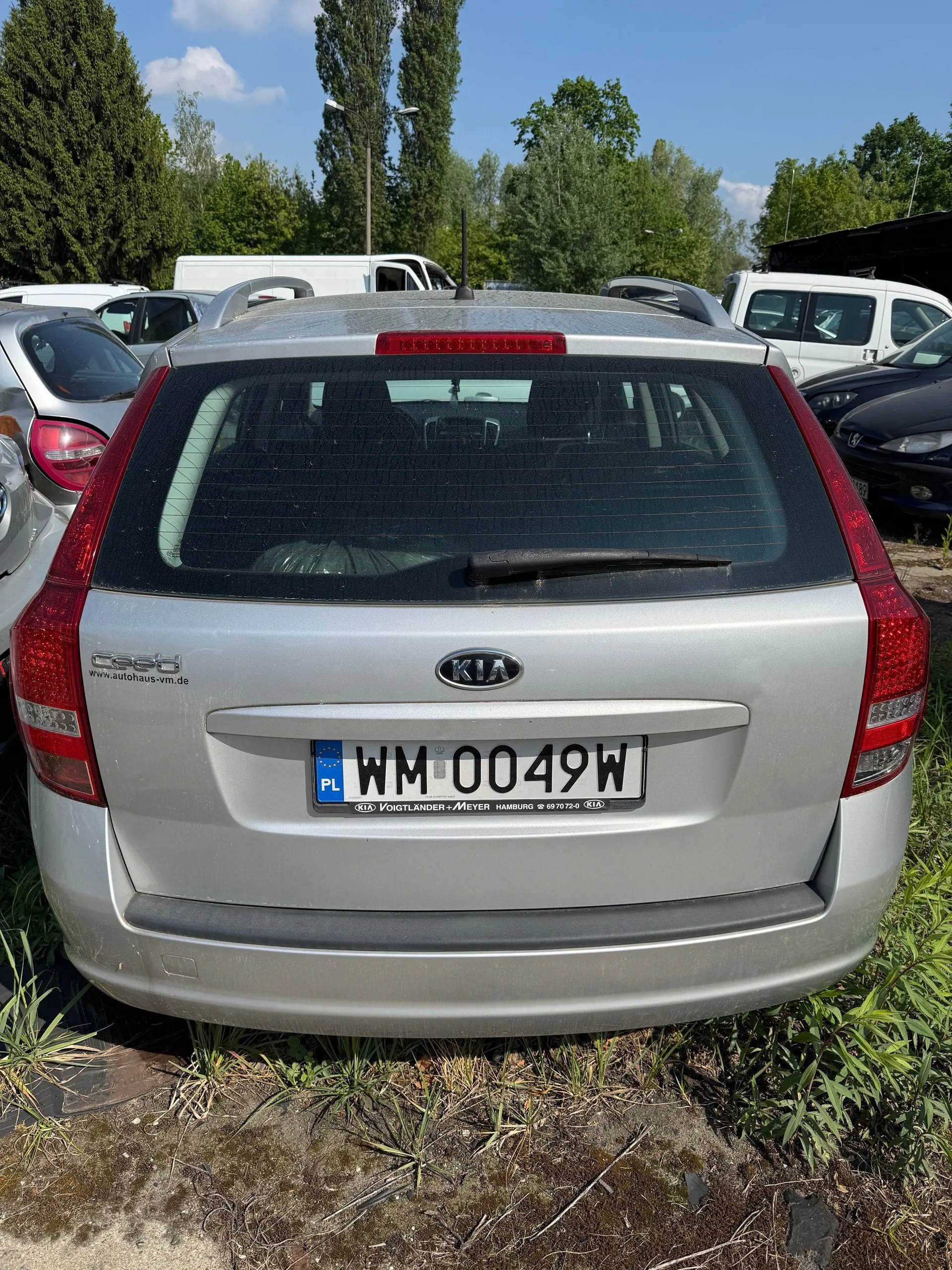 Tył srebrnego samochodu marki Kia Ceed z tablicą rejestracyjną WM 0049W (PL). Na tablicy widoczny napis "VOIGTLÄNDER+MEYER HAMBURG" oraz numer telefonu. Samochód stoi na trawie i ziemi, w tle inne pojazdy, drzewa i błękitne niebo z chmurami. Na tylnym szybie widoczny wycieraczka i czerwone światło. Po lewej stronie napis "www.autohaus-vm.de".
