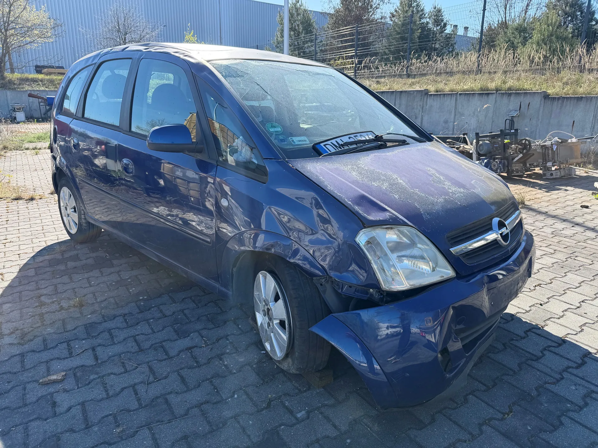 Niebieski samochód Opel z uszkodzonym przodem, w tym zdeformowaną maską i zderzakiem, zaparkowany na brukowanej powierzchni. Widoczna tablica rejestracyjna DML 2574. W tle betonowa ściana, maszyny przemysłowe, ogrodzenie i budynek z blachy falistej. Słoneczny dzień.
