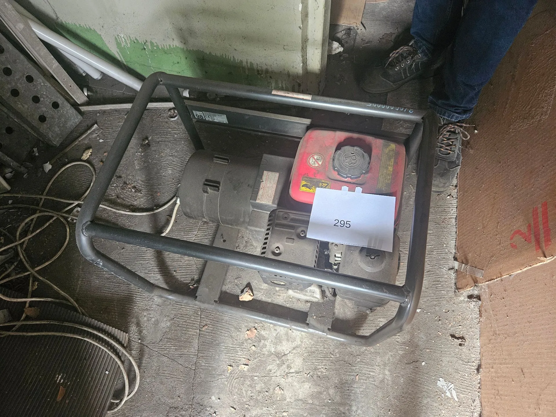 Zdjęcie z góry przedstawiające urządzenie techniczne w szarej metalowej ramie ochronnej. Wewnątrz ramy znajduje się szary silnik elektryczny oraz czerwony zbiornik z nakrętką. Na zbiorniku przyklejona jest biała kartka z napisem '295'. Urządzenie stoi na betonowej posadzce w pomieszczeniu gospodarczym. W prawym górnym rogu widoczne są nogi osoby w ciemnych spodniach i butach trekkingowych.