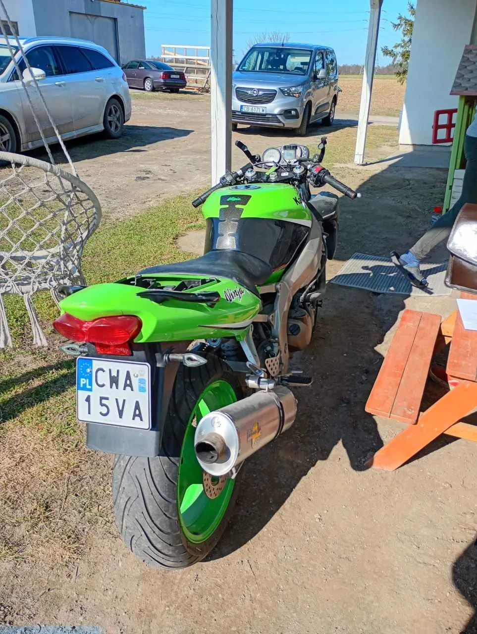Zdjęcie zielonego motocykla z tablicą rejestracyjną PL CWA 15VA, zaparkowanego na podwórku. W tle widoczne są samochody: srebrny SUV, ciemny sedan i srebrny van. Po lewej stronie znajduje się biała huśtawka w kształcie kosza, po prawej drewniany stół i ławka. Na pierwszym planie widoczne są elementy motocykla: czerwone światło tylnego, wydech, zielone felgi oraz czarna siedzisko. Tło stanowi budynek garażowy i otwarta przestrzeń z trawą.