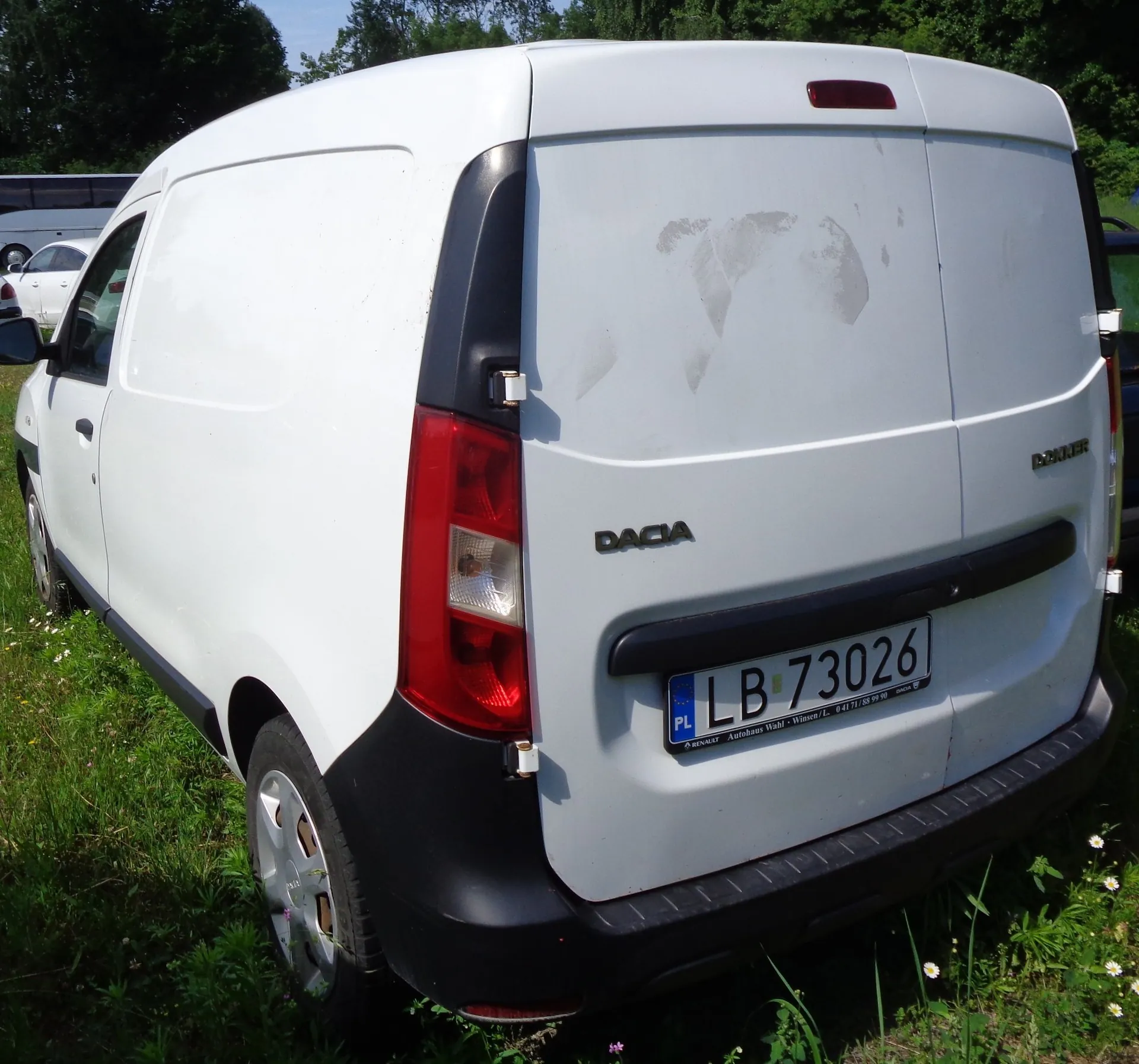 Biały samochód dostawczy marki Dacia widziany od tyłu i z boku, zaparkowany na trawie. Na tylnych drzwiach widnieje napis DACIA oraz tablica rejestracyjna o numerze LB 73026 z oznaczeniem PL. Na białym lakierze tylnych drzwi widoczne są zatarcia i ślady zużycia.