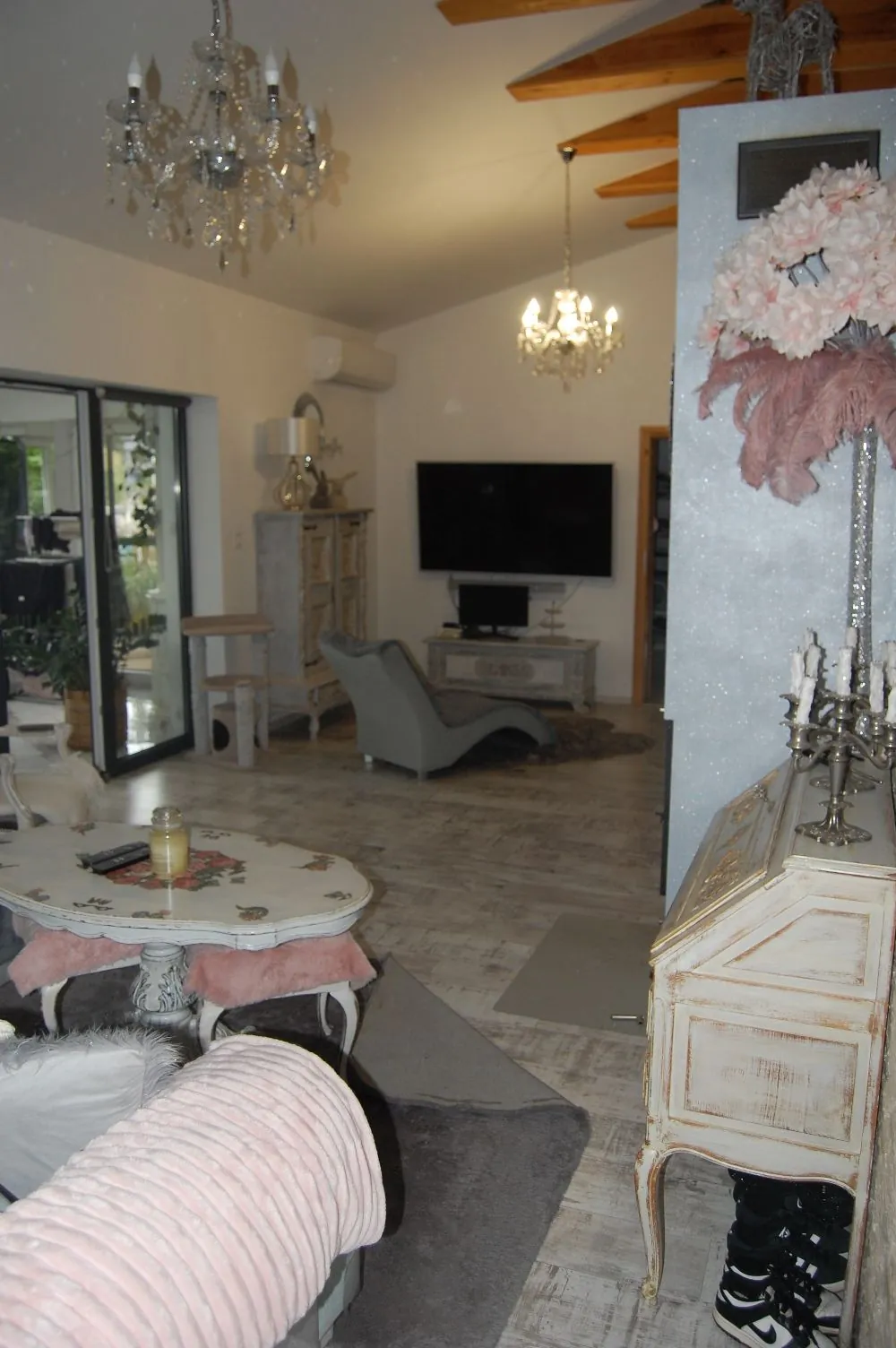Wnętrze jasnego salonu w stylu vintage. W pomieszczeniu znajduje się biały, zdobiony stolik kawowy z różowymi poduszkami, szary fotel, biała komoda oraz telewizor zamontowany na ścianie. Z sufitu zwisają dwa kryształowe żyrandole. Po prawej stronie widoczna jest biała szafka z dekoracyjnym kwiatem i świecznikami na górze. Podłoga wykonana jest z jasnych paneli drewnianych.