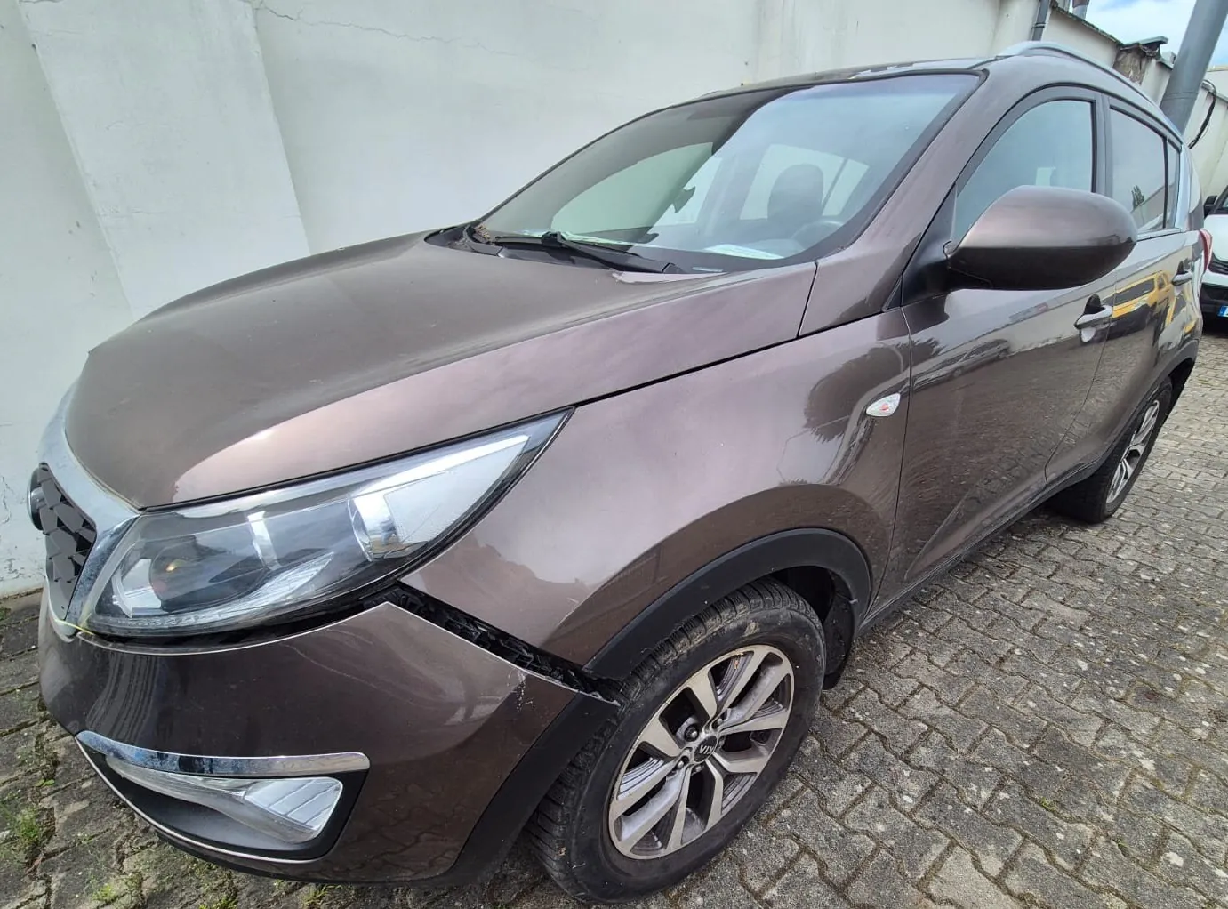Brązowy samochód typu SUV zaparkowany na kostce brukowej obok białej ściany. Pojazd jest widoczny z przodu i z boku, z wyraźnym uszkodzeniem zderzaka przedniego w miejscu łączenia z błotnikiem po stronie kierowcy.
