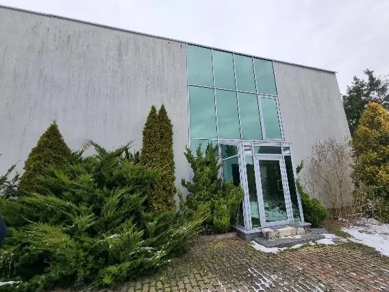 Budynek o szarej betonowej elewacji z dużą sekcją szklanych okien i drzwi wejściowych z metalowym profilem. Przed budynkiem rosną zielone krzewy i drzewa iglaste, częściowo pokryte śniegiem. Nawierzchnia z ceglanego brukowania z resztkami śniegu. Niebo jest zachmurzone.