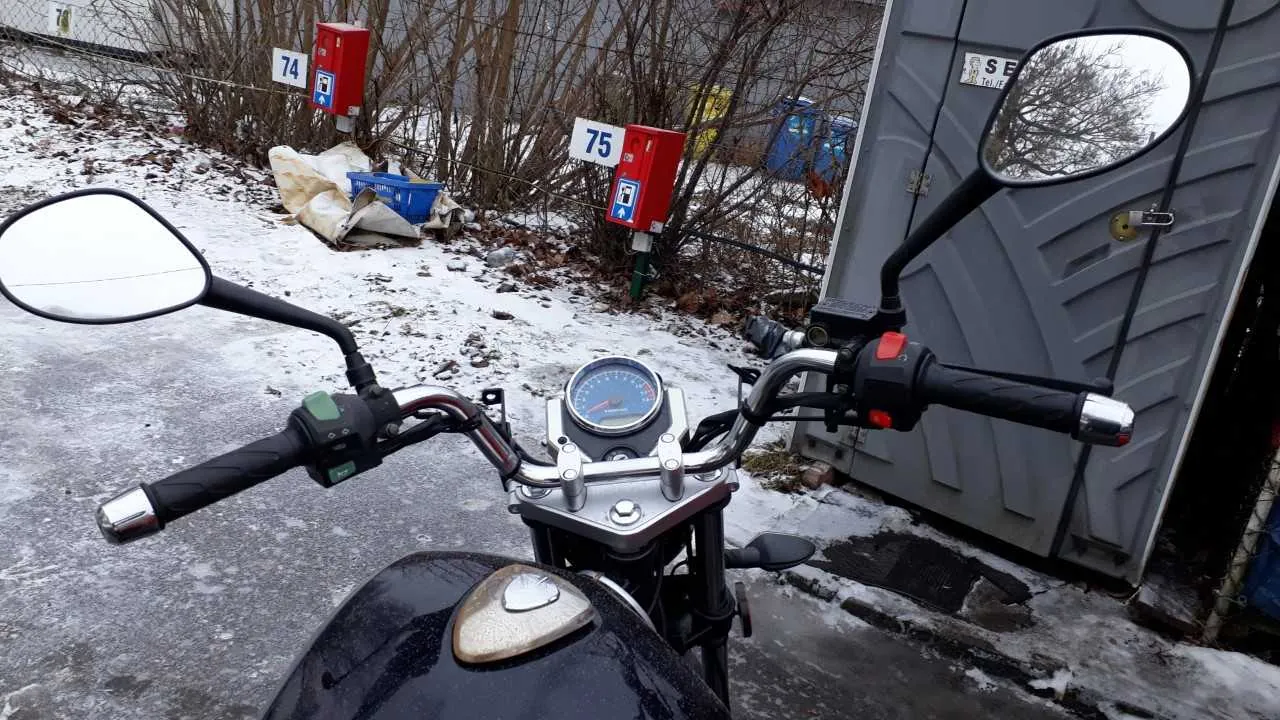 Widok z perspektywy kierowcy na czarny motocykl z widocznymi kierownicą, licznikiem prędkości i lusterkami bocznymi. Na tle śnieżna powierzchnia, krzaki, dwa czerwone pudełka z białymi tabliczkami oznaczonymi numerami 74 i 75 oraz niebieskimi symbolami parkingu. Po prawej stronie szara przenośna toaleta z widocznym zamkiem i częściowo czytelnym napisem. Na ziemi leżą niebieskie kosze i worki. Tło stanowi ogrodzenie i budynki.