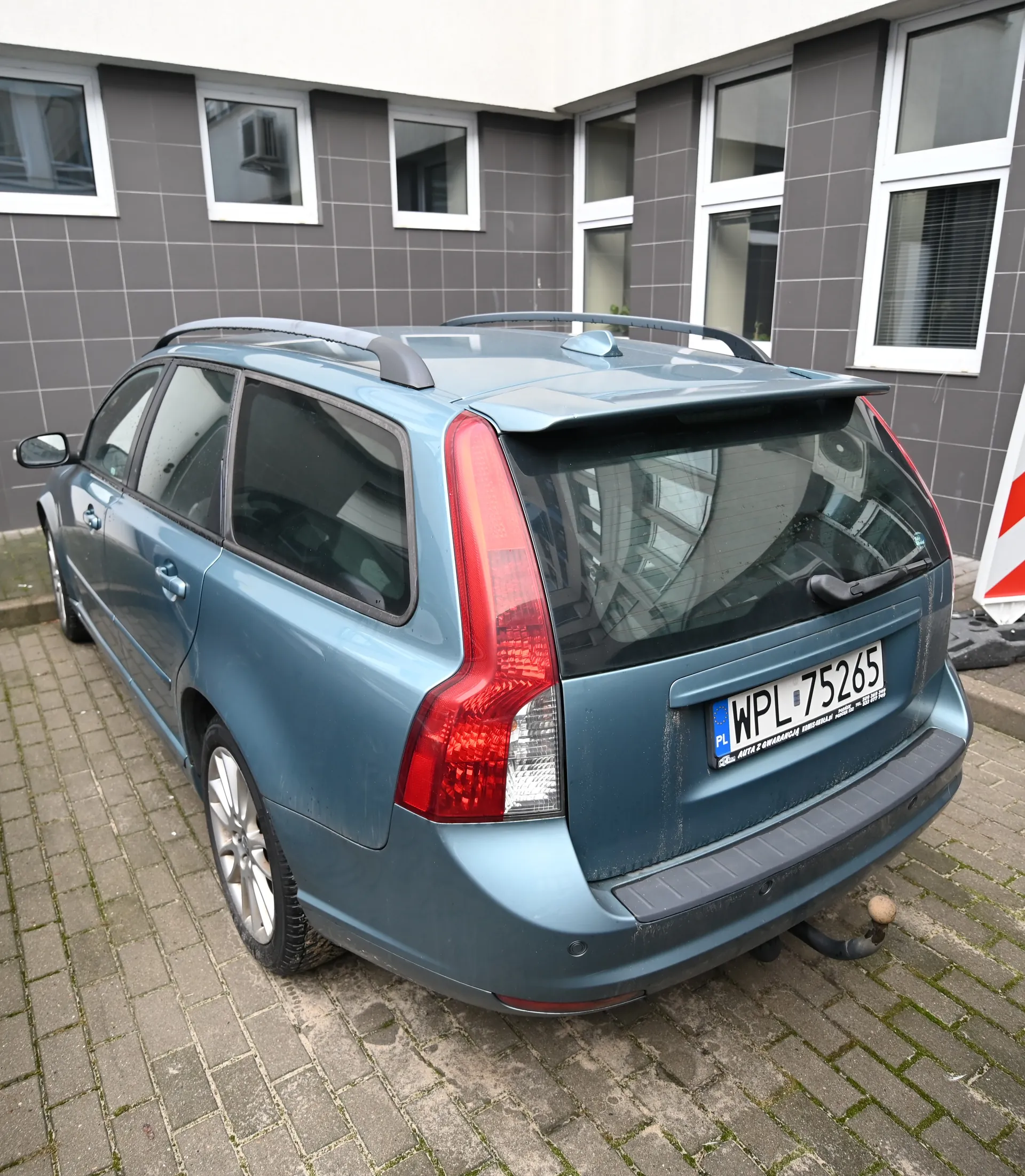Niebieski samochód typu kombi z dachowym bagażnikiem i spojlerem tylnym. Tablica rejestracyjna WPL 75265. Zaparkowany na brukowanej powierzchni przed budynkiem z szarymi płytkami i białymi oknami. Widoczny czerwono-biały znak ostrzegawczy.