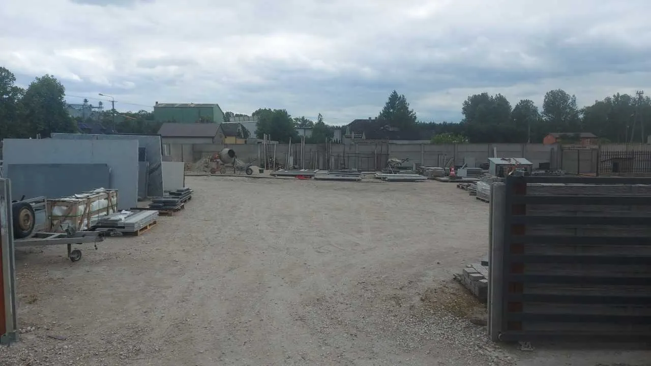 Budowa z otwartym terenem o podłożu z żwiru i ziemi. Po lewej stronie widoczne są materiały budowlane, w tym betonowe płyty, przyczepa z kołami i palety. W tle stoją budynki, drzewa oraz betoniarka. Po prawej stronie znajduje się drewniana konstrukcja. Niebo jest zachmurzone. Brak czytelnego tekstu.