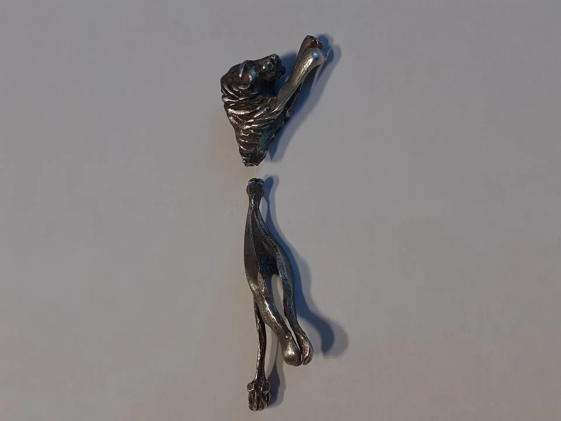 Srebrzysta, metalowa figurka zwierzęcia, prawdopodobnie psa, która została przełamana na dwie części. Górna część przedstawia głowę i przednią łapę, dolna część przedstawia tylne łapy. Obie części leżą na gładkim, jasnoszarym tle.