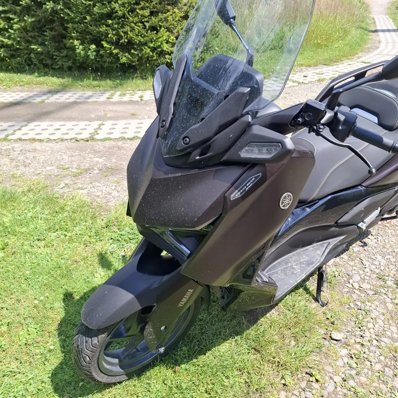 Czarny skuter marki Yamaha zaparkowany na trawie i żwirze. Widoczna jest przednia część pojazdu z wysoką, przezroczystą szybą ochronną, reflektorami oraz napisem YAMAHA na przednim błotniku. W tle widać zielone krzewy i fragment betonowej ścieżki.