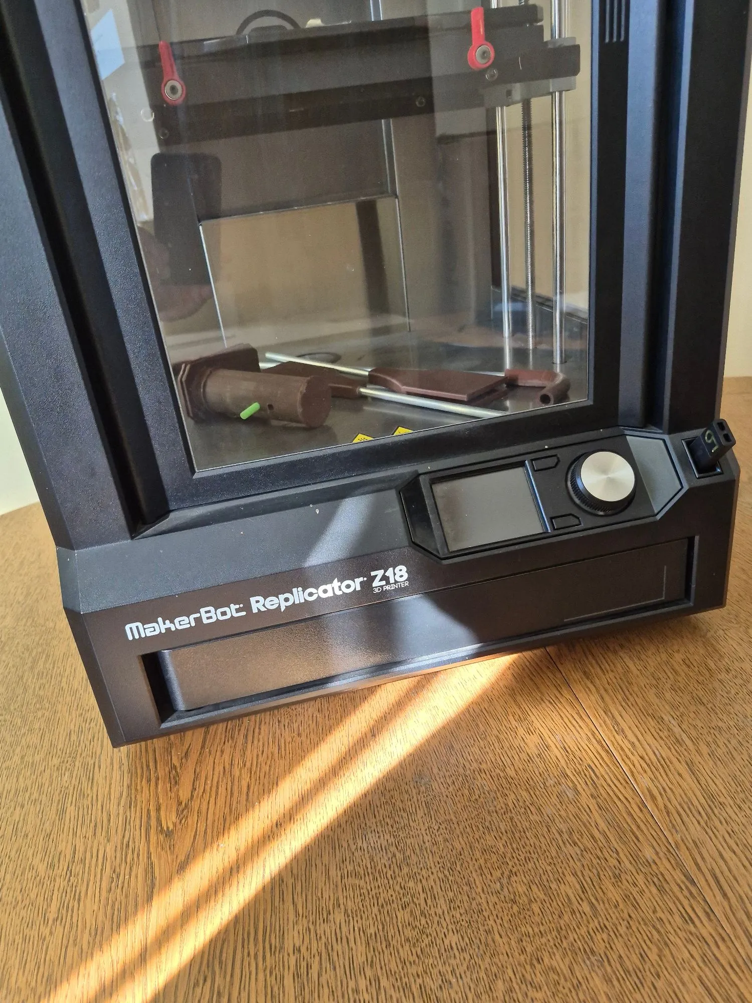 Zdjęcie przedstawiające fragment czarnej drukarki 3D MakerBot Replicator Z18. Widoczna jest zamknięta komora z przezroczystą szybą, wewnątrz której znajdują się brązowe wydruki z tworzywa. Na przednim panelu urządzenia znajduje się ekran LCD, pokrętło oraz napis: 'MakerBot Replicator Z18 3D PRINTER'. Urządzenie stoi na drewnianym blacie, na który pada pasmo słonecznego światła.