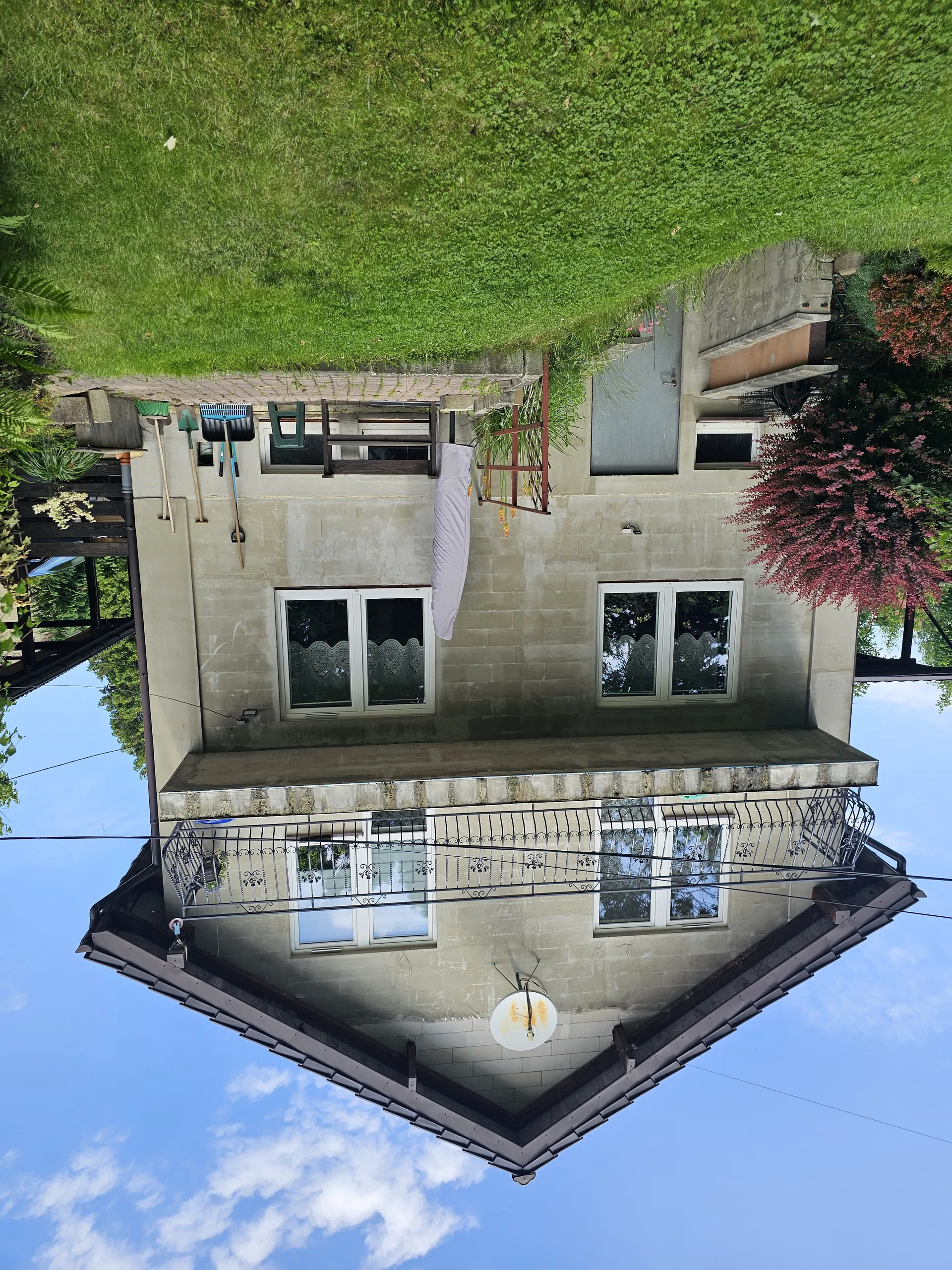 Dwupiętrowy dom o beżowej elewacji z dachem dwuspadowym. Na elewacji widoczne są okna z białymi ramami, balkon z czarnym metalowym ogrodzeniem oraz biała antena satelitarna. Przy ścianie domu stoją narzędzia ogrodnicze, a na podwórku znajduje się zielona trawa, czerwony krzew i składany parasol. W tle błękitne niebo z białymi chmurami.