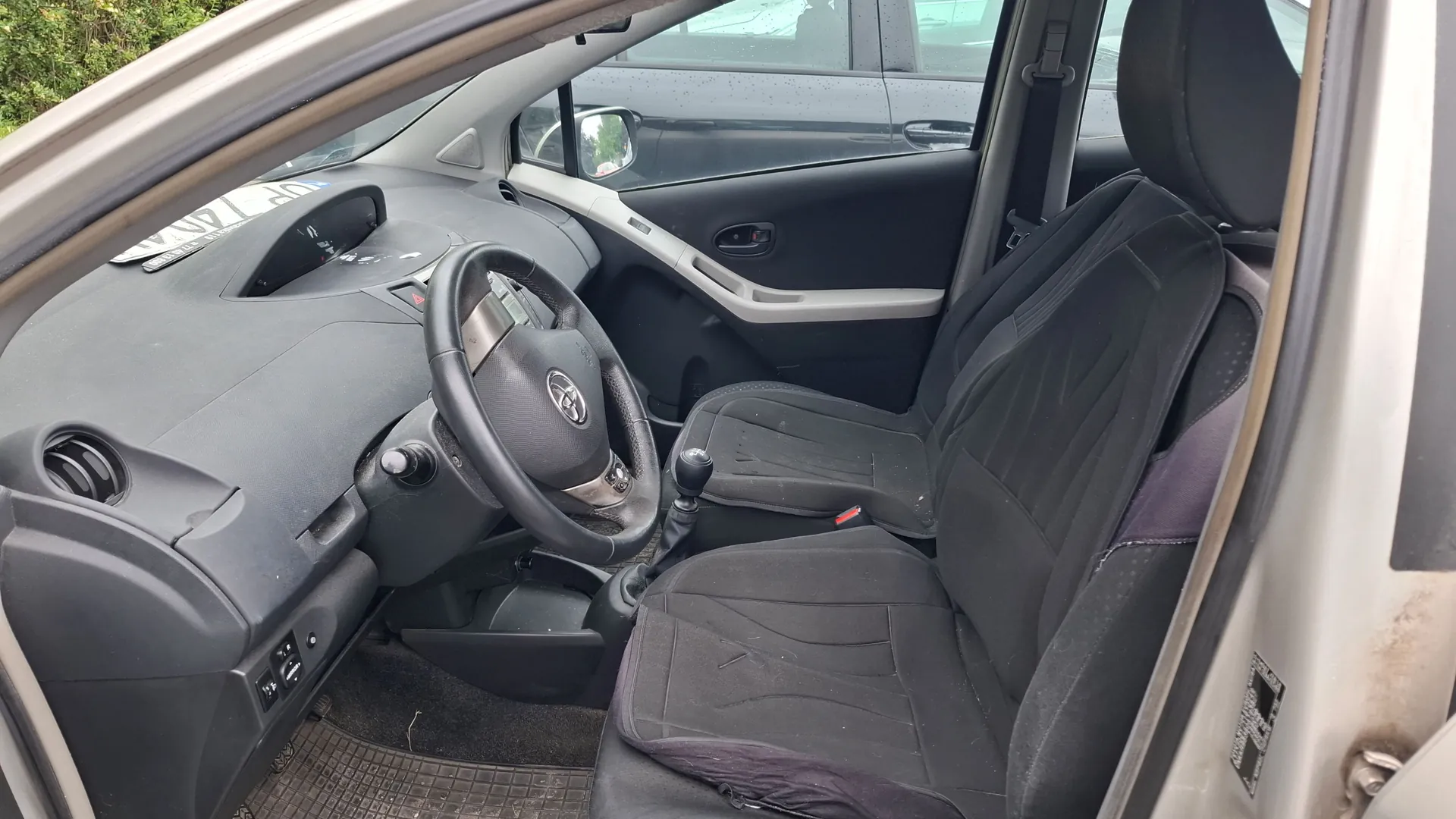 Wnętrze samochodu widziane z otwartych drzwi pasażera. Widoczna jest czarna tapicerka foteli, kierownica z logo Toyoty, deska rozdzielcza, lewarek zmiany biegów oraz fragmenty srebrnych wykończeń wewnątrz kabiny.