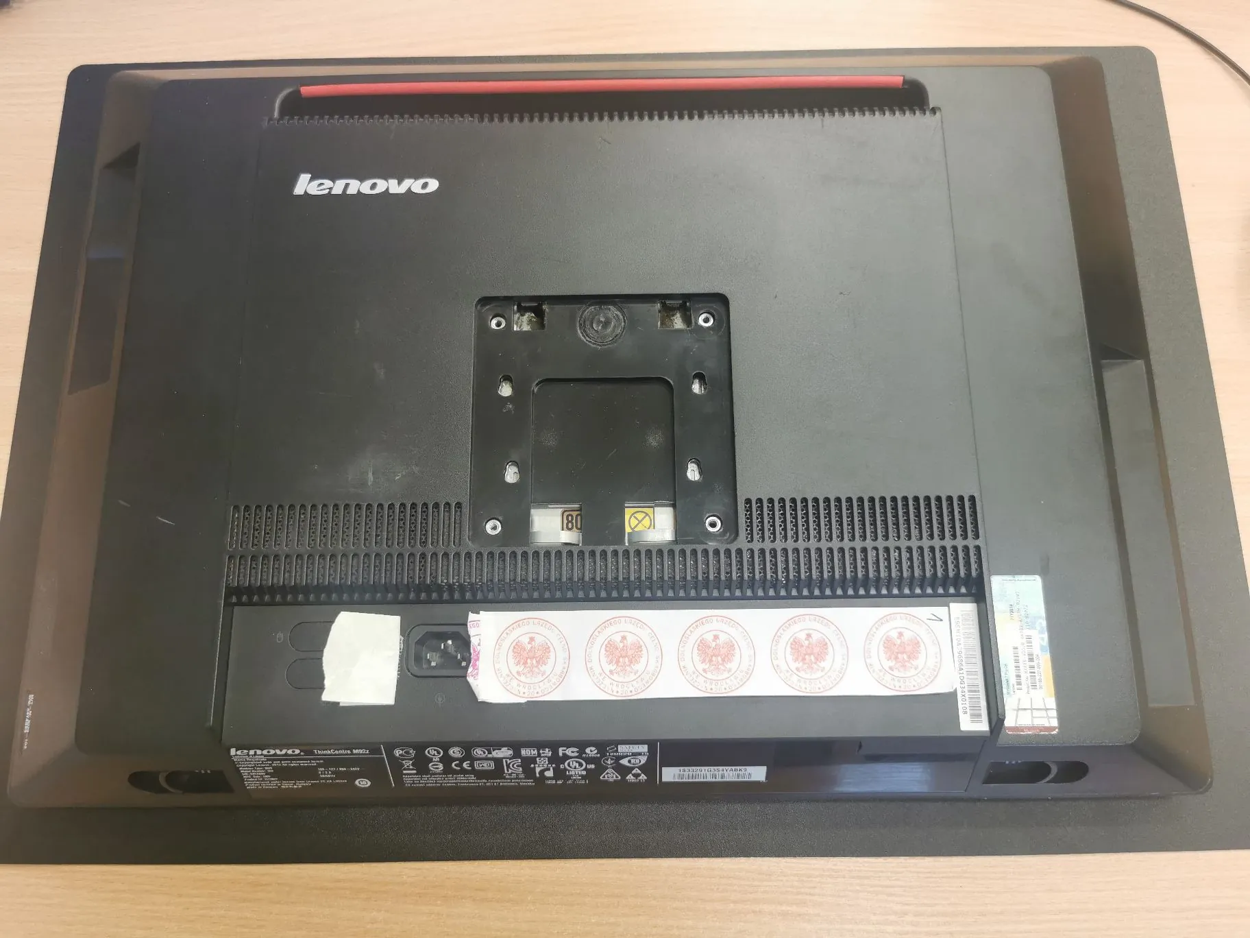 Zdjęcie przedstawiające tylny panel czarnego monitora marki Lenovo. Na górze widoczne jest białe logo 'lenovo'. W centralnej części znajduje się uchwyt VESA do montażu na ścianie. Poniżej znajdują się otwory wentylacyjne oraz naklejki z informacjami technicznymi i certyfikatami. Na dole widnieje tabliczka znamionowa z tekstem 'lenovo ThinkCentre M90t' oraz symbolami takimi jak CE, FCC i inne oznaczenia regulacyjne.