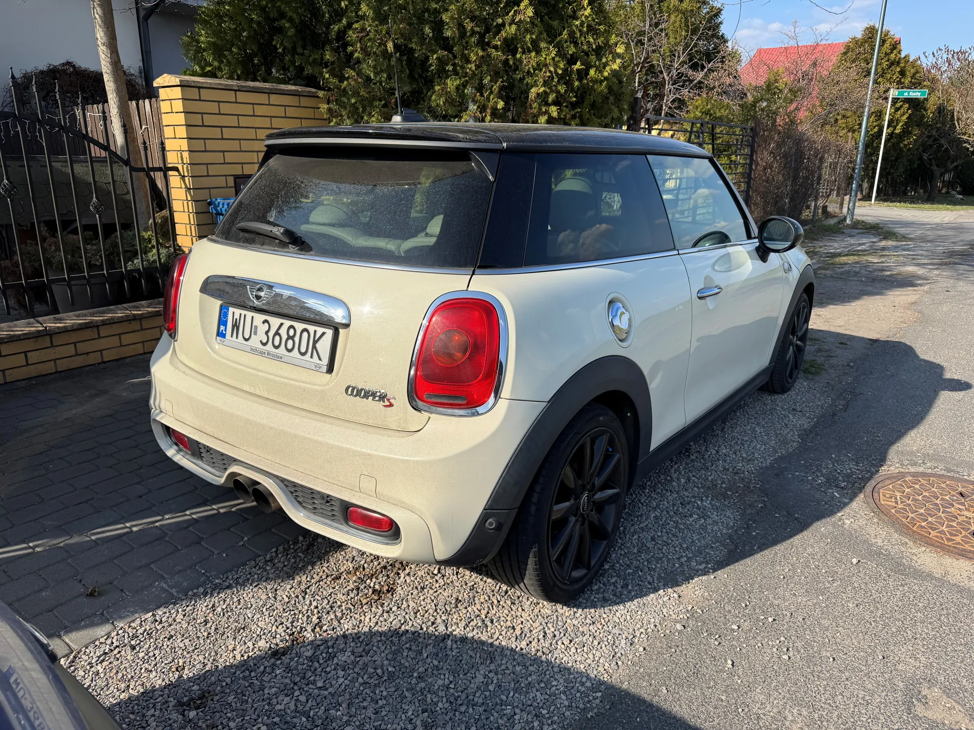 Biały samochód Mini Cooper S zaparkowany na żwirowym i brukowanym obszarze. Widok z tyłu z tablicą rejestracyjną WU 3680K. W tle żółta ceglana ściana z czarnym metalowym ogrodzeniem, drzewa, dom z czerwonym dachem oraz znak uliczny. Słoneczna pogoda z jasnym niebem.