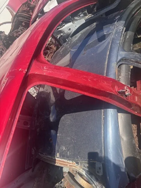 Zbliżenie na uszkodzone fragmenty samochodów w stanie wraku. Widoczne są czerwone i niebieskie elementy karoserii, części mechaniczne oraz śladami korozji i brudu. W tle fragmenty innych zniszczonych pojazdów. Brak czytelnego tekstu.
