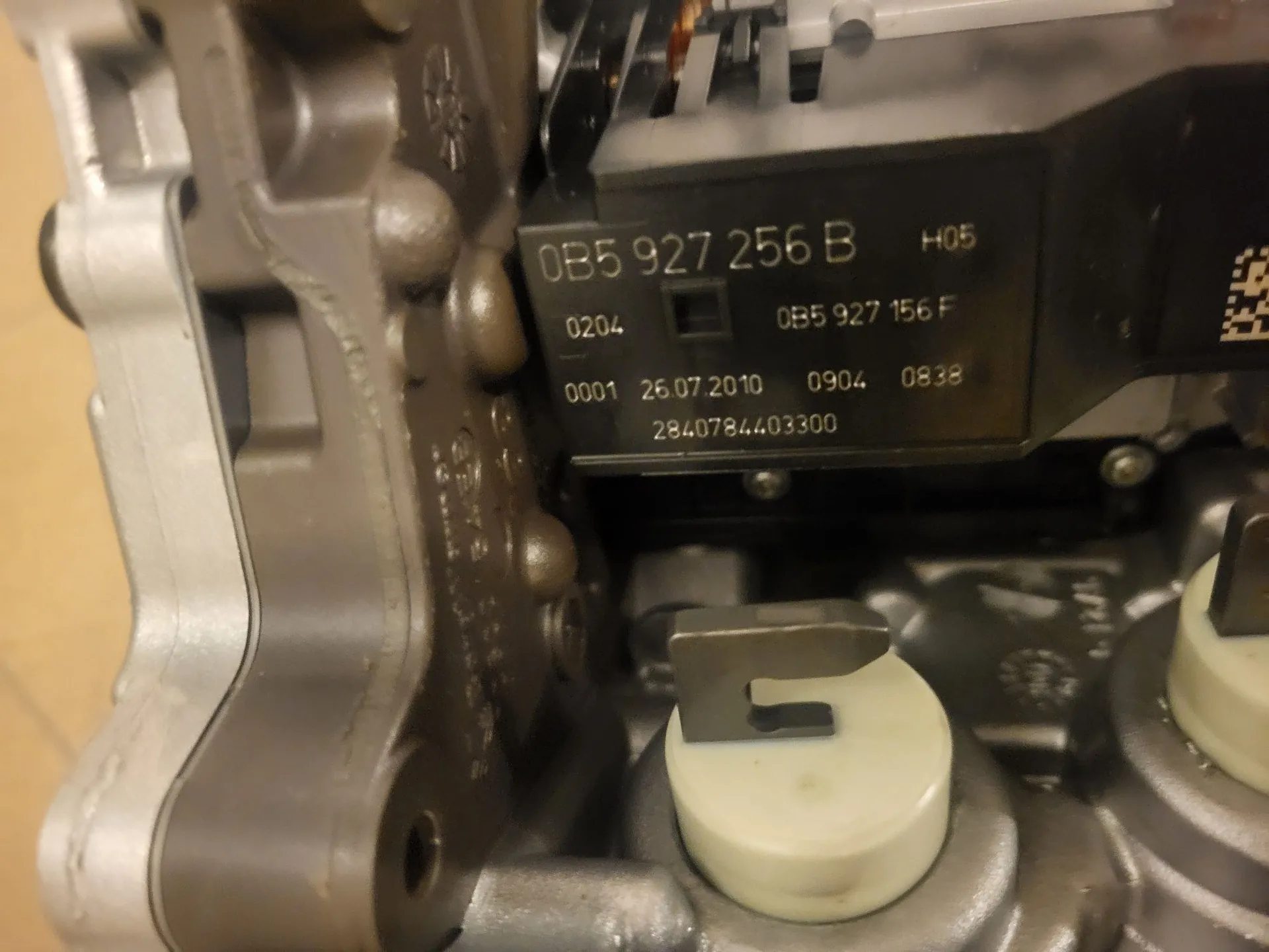 Zbliżenie na czarną tabliczkę znamionową przymocowaną do metalowej części silnika. Na tabliczce znajduje się biały tekst: '0B5 927 256 B H05', poniżej '0204 0B5 927 156 F', następnie '0001 26.07.2010 0904 0838' oraz na dole numer '2840784403300'. Po lewej stronie widoczny jest odlew metalowy z wypustkami, a poniżej biały plastikowy element z metalowym zaczepem.