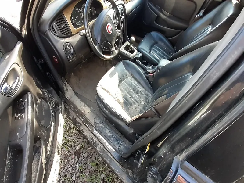 Wnętrze samochodu z otwartą kierowcą drzwiami. Widoczne czarne skórzane fotele w stanie zużycia, kierownica z logo, deska rozdzielcza z wskaźnikami i wentylacją. Podłoga pokryta brudem i śmieciem. Z zewnątrz widoczny żwir i trawa.