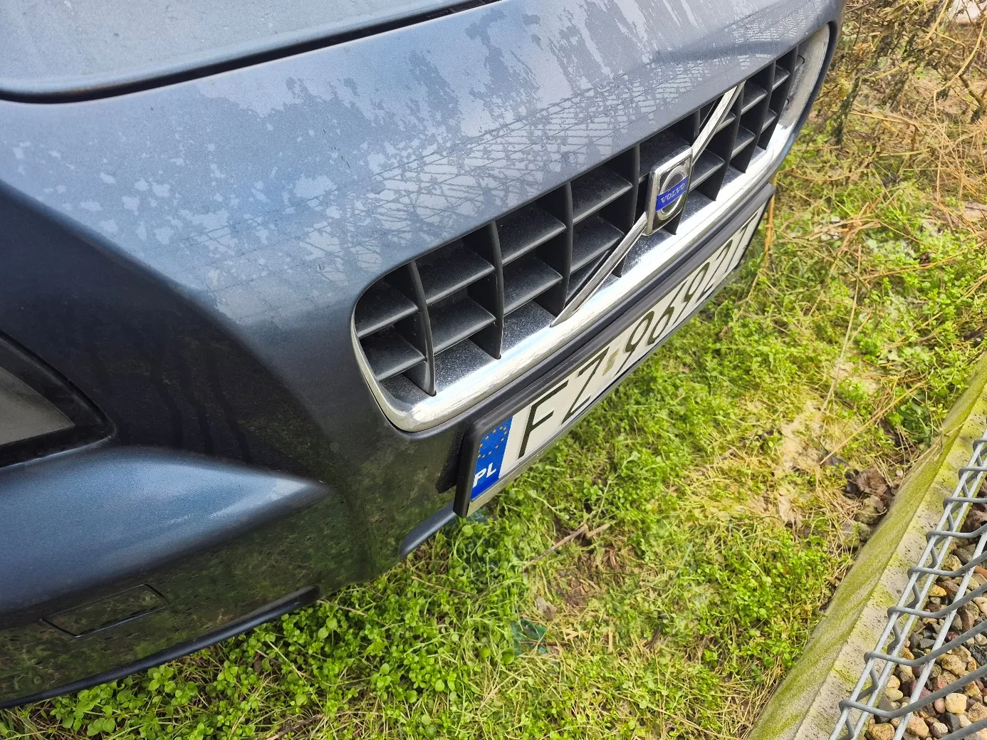 Przód ciemnoszarego samochodu marki Volvo z widoczną atrapą chłodnicy i logo. Tablica rejestracyjna z polskim kodem 'PL' i numerem 'FZ 9692'. Samochód zaparkowany na trawie z suchymi roślinami i metalowym ogrodzeniem po prawej stronie. Na masce widoczne białe plamy.