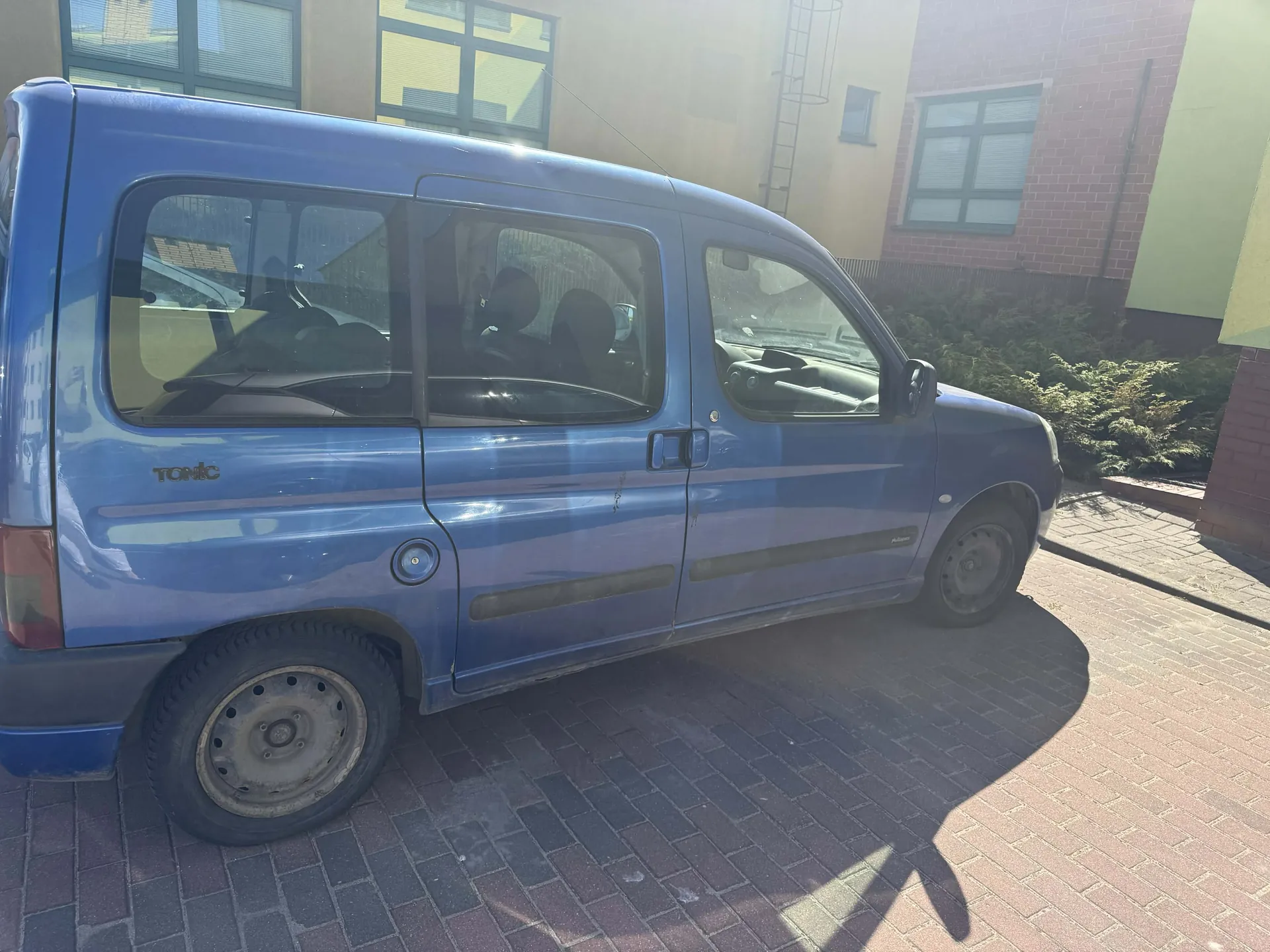 Niebieski samochód dostawczy marki Tonic zaparkowany na brukowanej powierzchni. Widoczne są boczne drzwi, okna z siedzeniami wewnątrz oraz napis "Tonic" na tylnej części pojazdu. W tle budynki z oknami, drabina pożarna i krzewy. Cień samochodu pada na bruk. Opony mają metalowe felgi, a nadwozie posiada czarne listwy dekoracyjne.