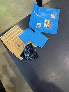 Na ciemnej powierzchni leżą niebieskie foldera, z których jeden zawiera zdjęcie osoby, czarna torba plastikowa oraz kartonowe pudełko z poziomymi paskami. W lewym dolnym rogu widoczna jest betonowa podłoga.