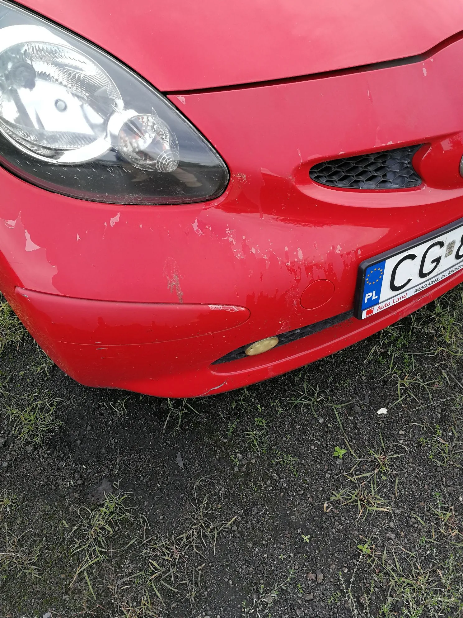 Zbliżenie na przedni prawy róg czerwonego samochodu. Widoczny jest reflektor, fragment maski oraz zderzaka z licznymi zarysowaniami i odpryskami lakieru. W prawym dolnym rogu widoczny jest fragment tablicy rejestracyjnej z symbolem PL oraz literami CG, a poniżej napis Auto Land oraz WENCLAWIEK PL KROSNO.