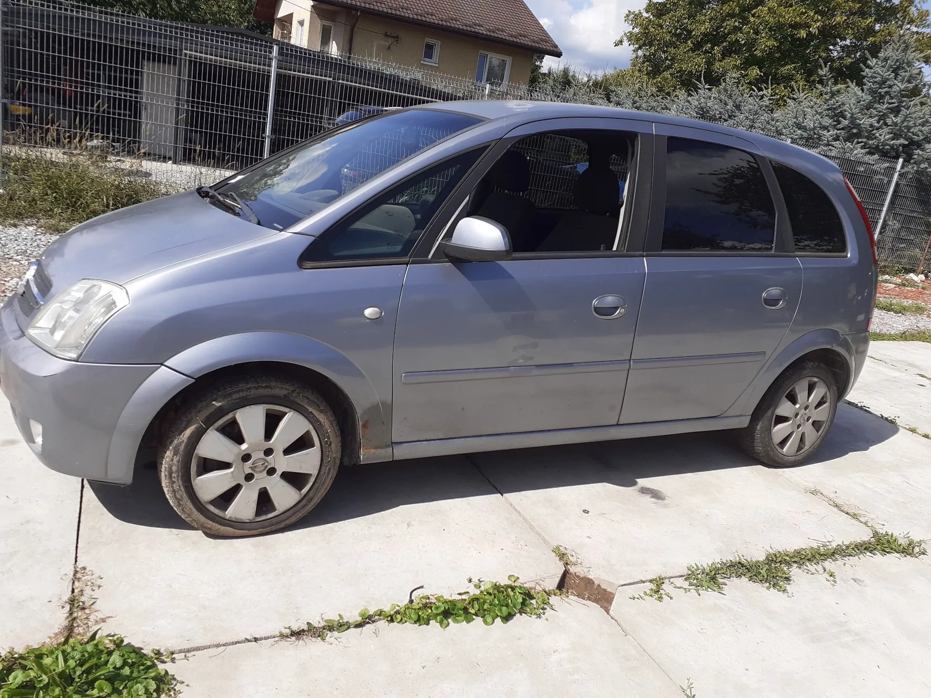 Niebieski samochód osobowy typu hatchback zaparkowany bokiem na betonowym podjeździe. W tle widoczny jest metalowy płot, dom oraz zielone drzewa i krzewy.
