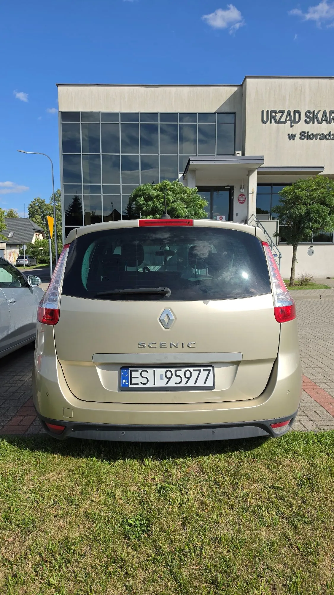 Z tyłu złoty samochód marki Renault Scenic z polską tablicą rejestracyjną ESI 95977. Auto zaparkowane jest na trawie przed nowoczesnym budynkiem z dużymi przeszklonymi ścianami. Na budynku widnieje napis „URZĄD SKARBOWY w Sieradzu”. W tle widać niebieskie niebo z kilkoma białymi chmurami.