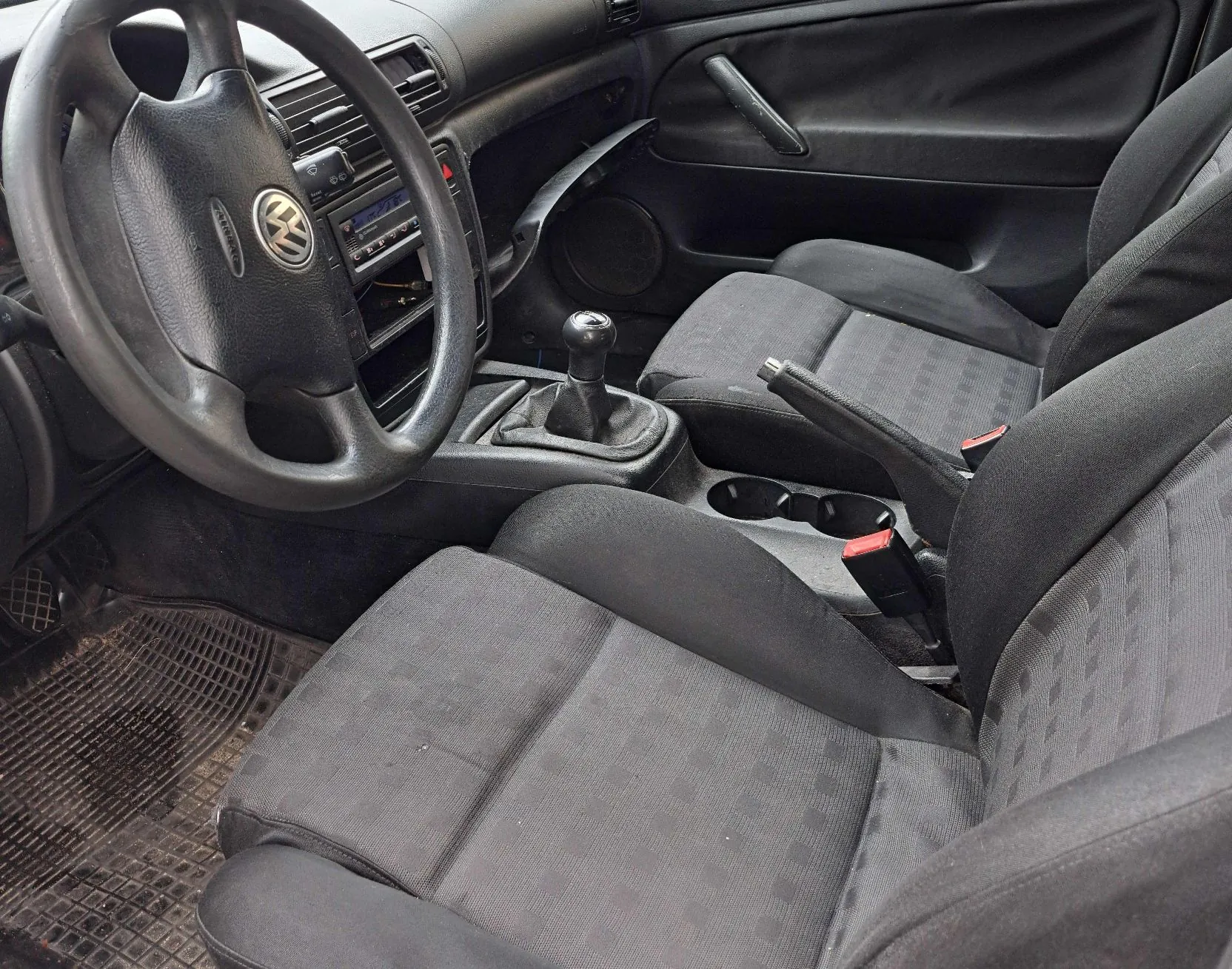 Wnętrze samochodu z perspektywy przedniego fotela pasażera. Widoczna jest czarna tapicerka materiałowa foteli, kierownica z logo Volkswagena, deska rozdzielcza z radiem, lewarek zmiany biegów oraz podłokietnik w konsoli środkowej.